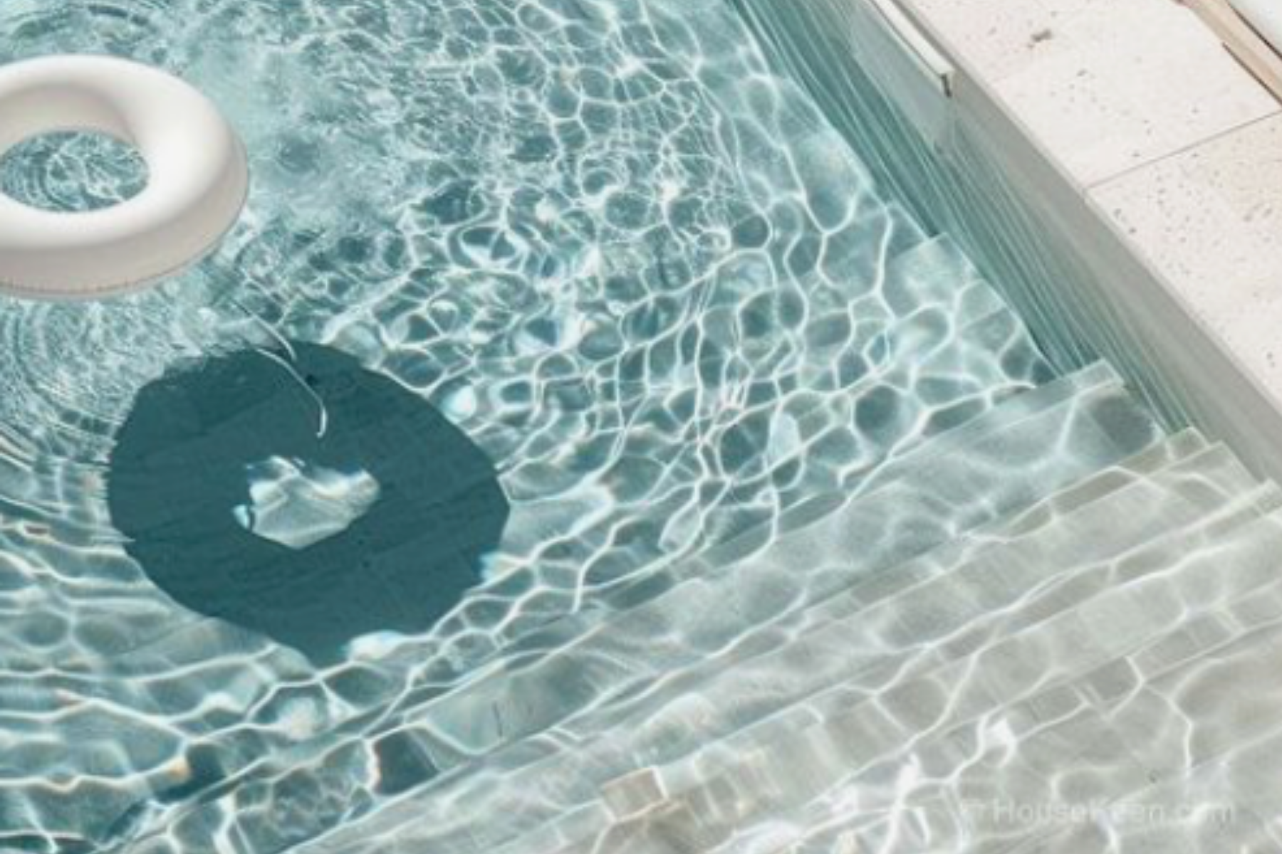 An inflatable white pool float ring with a cut-out handle floating on a clear blue swimming pool with sunlight reflections.