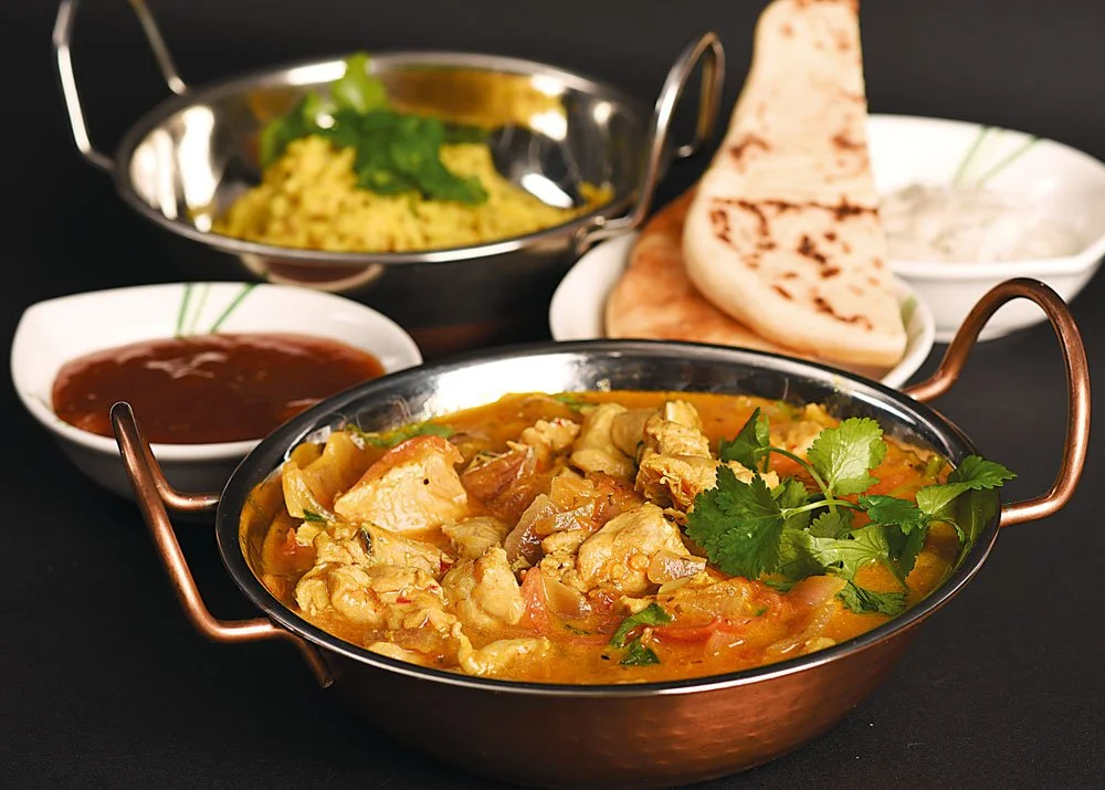 Indian curry dish garnished with cilantro, served with naan bread, rice, and side sauces.