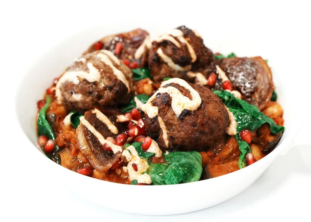 A white bowl of Indian-style meatballs with spices, topped with a drizzle of cream or yogurt, served over a bed of vegetables including spinach and onions, garnished with pomegranate seeds.