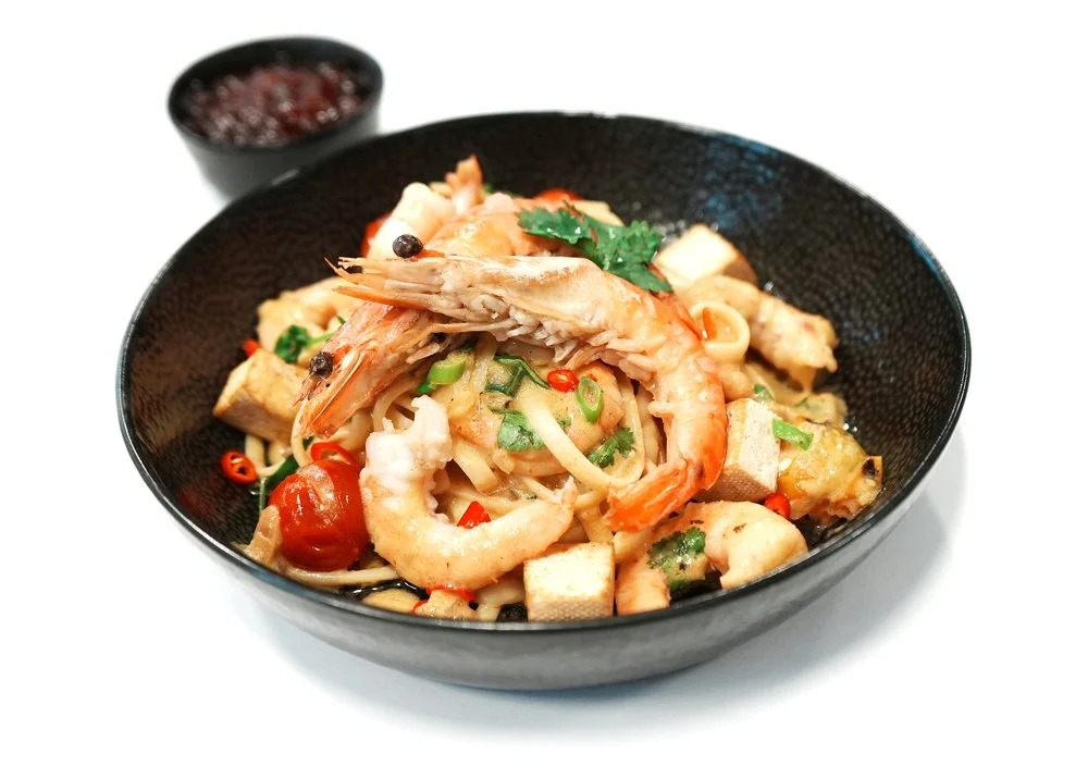 Bowl of seafood pasta with shrimp, cherry tomatoes, green onions, and cilantro, served with a small side dish of soy sauce or dipping sauce