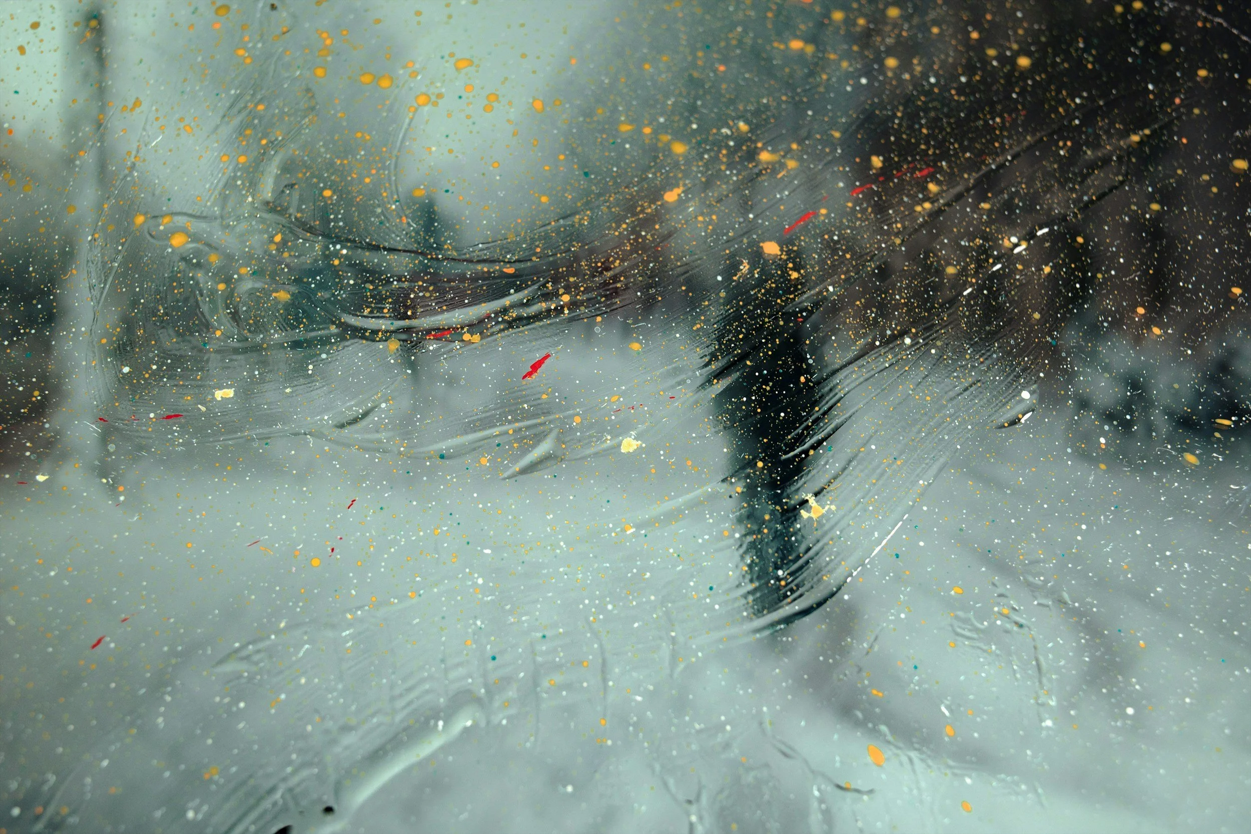 Close-up of a foggy window with water droplets and paint splatters in gold, red, and other colors.