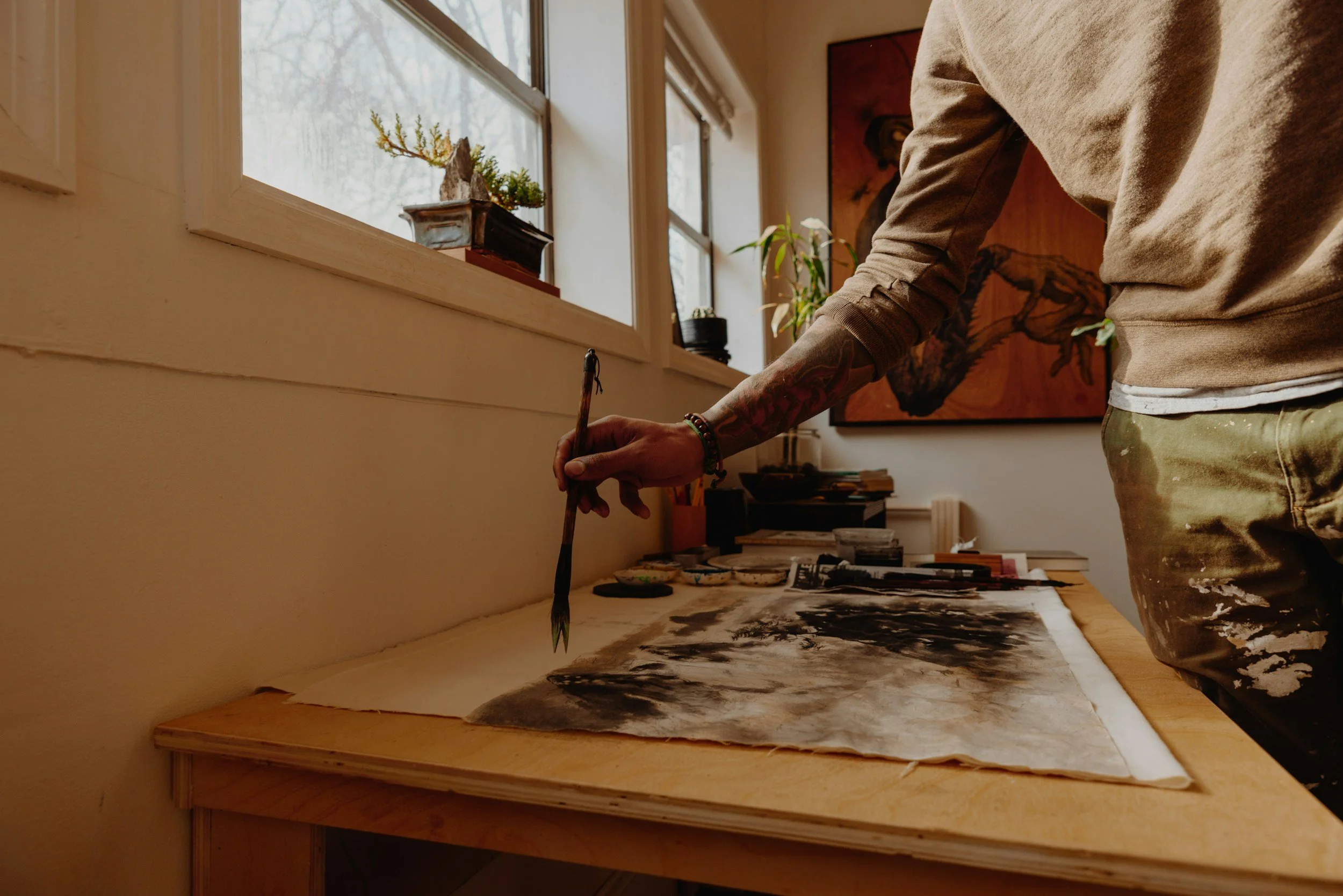 An artist working on a painting at a wooden table in a cozy art studio, with plants and artwork in the background.