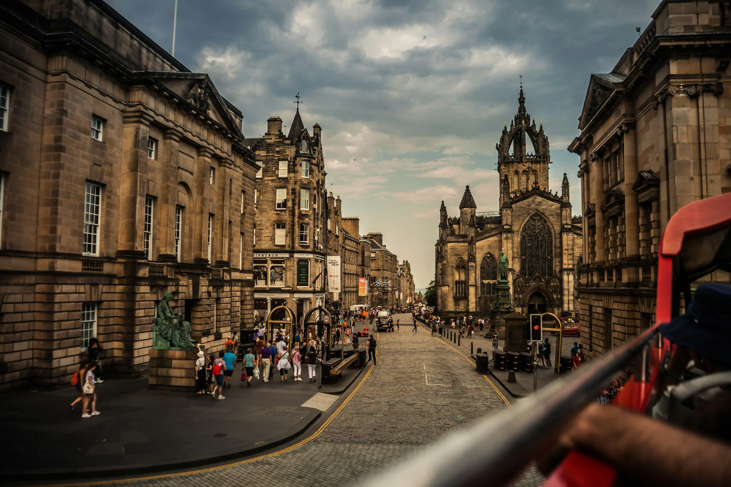 A busy city street with historic architecture, people walking, and a church with a tall spire in the background, under cloudy skies.