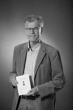 Black and white photo of a man in glasses wearing a blazer, holding a booklet or magazine.