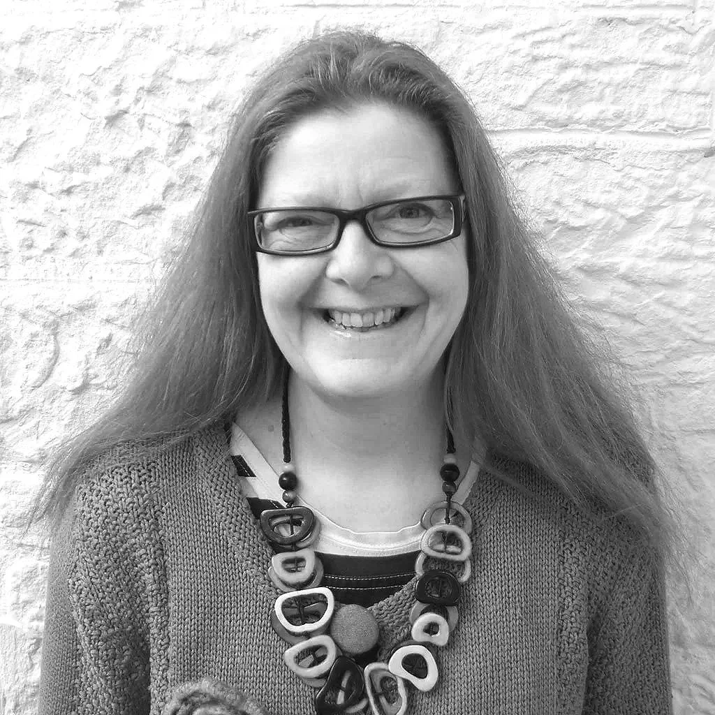 A woman with long hair and glasses smiling in front of a textured wall, wearing a sweater and a bold statement necklace.
