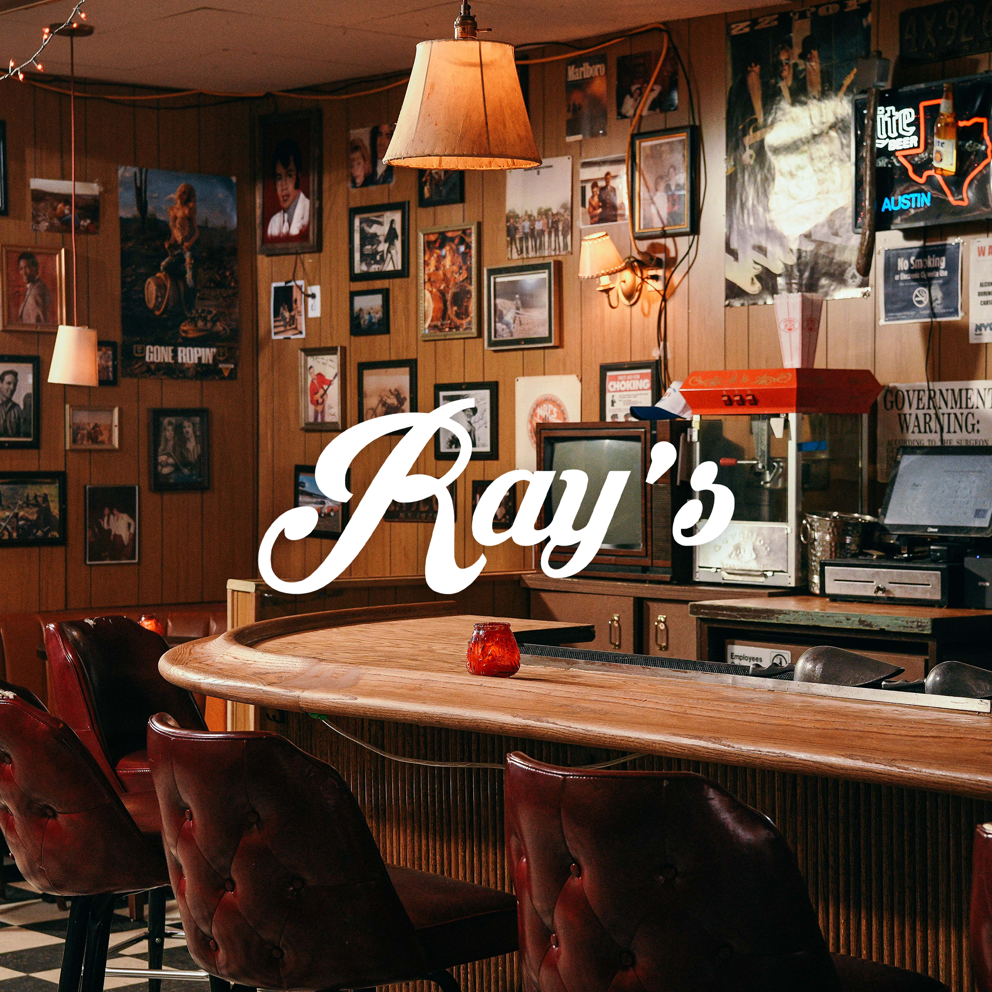 Cozy bar with wooden walls decorated with various posters and photos, a curved wooden bar counter with red leather chairs, hanging lamps, a small red candle holder on the counter, and vintage decor including a popcorn machine and TVs.