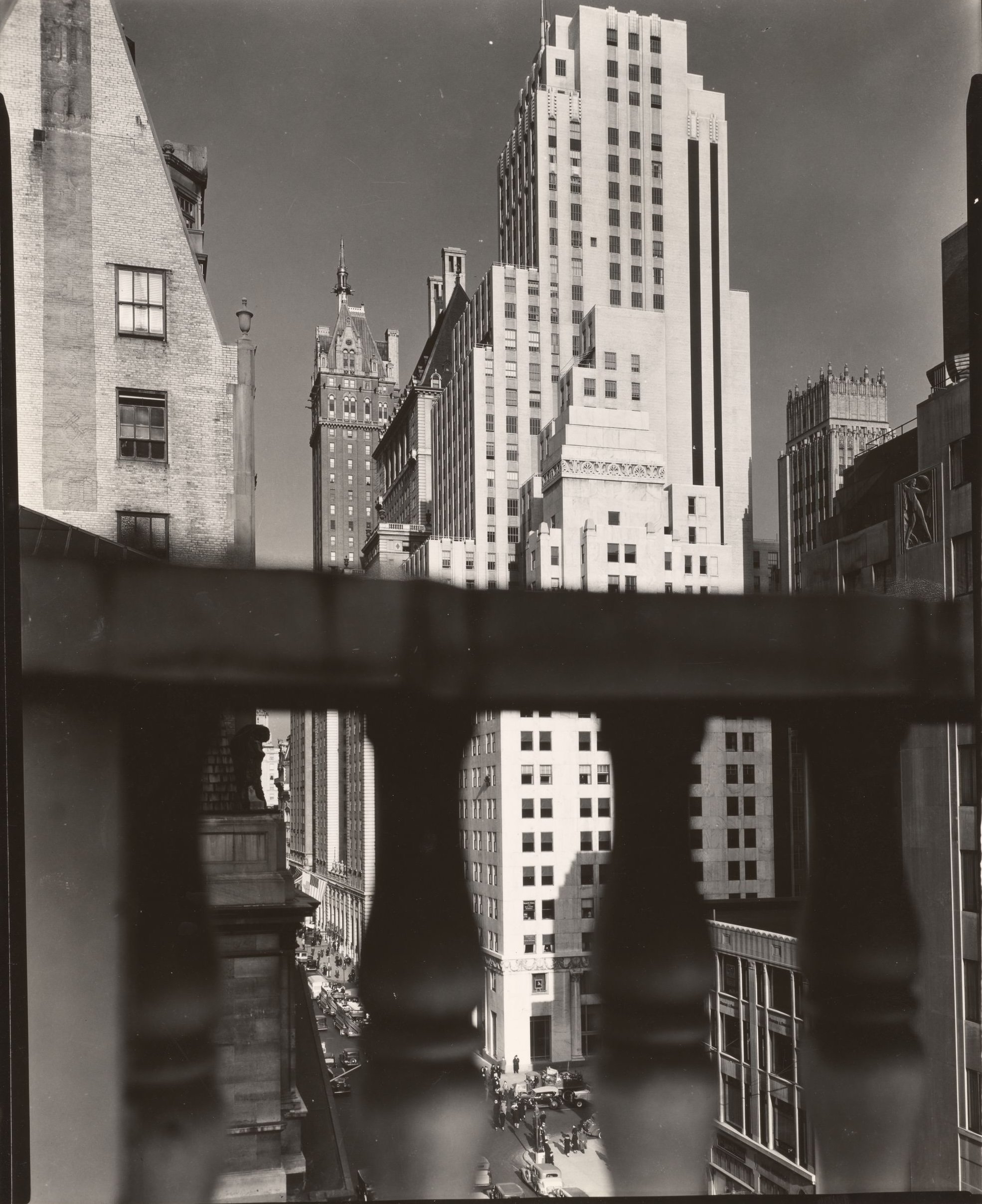 Vintage black and white photograph of a Manhattan cityscape viewed from a balcony with a railing in the foreground. Tall skyscrapers and historic buildings line the streets below.