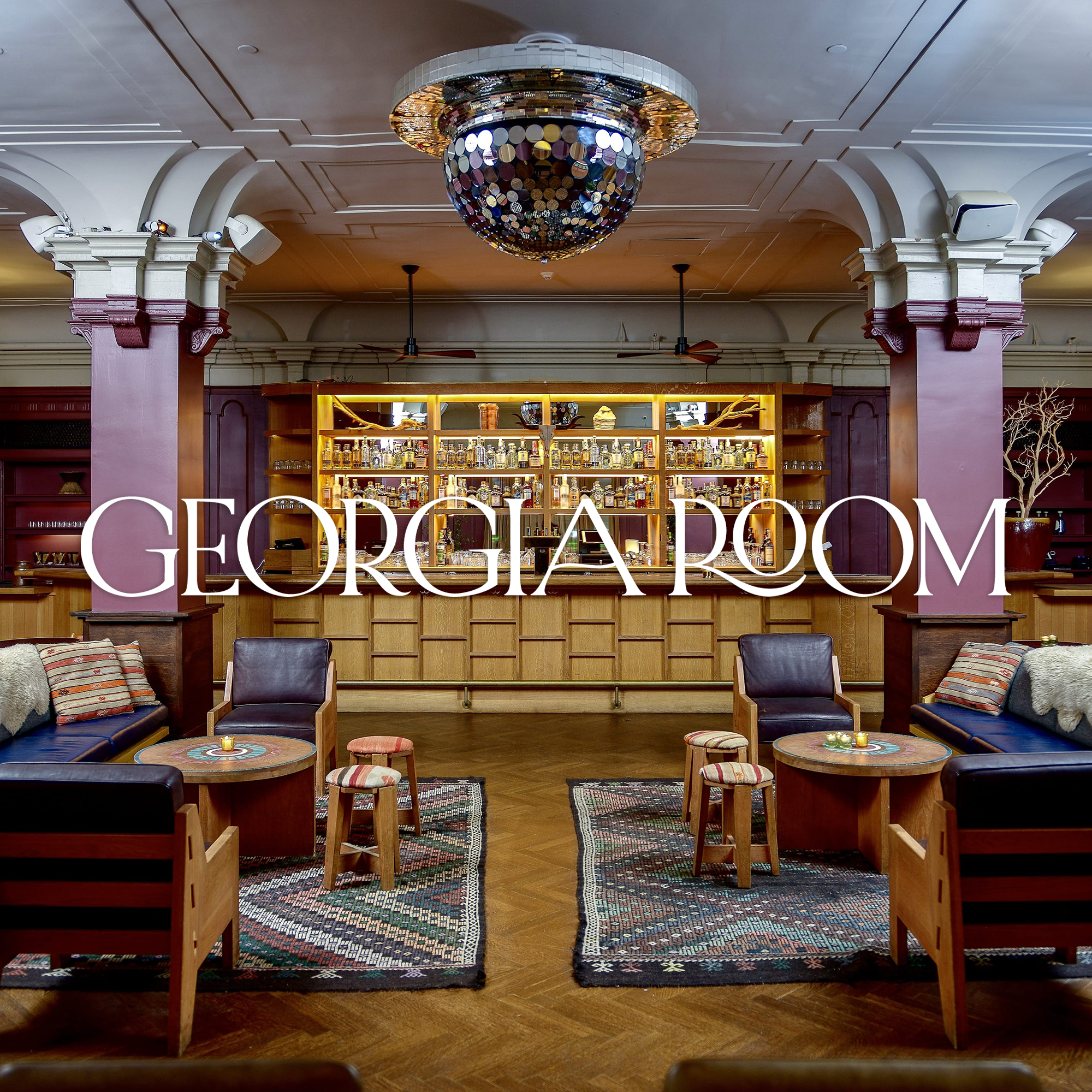 Interior of the Georgia Room, featuring a bar with bottles on shelves, seating with plush chairs and small tables, a chandelier above, and decorative elements like a large potted plant and colorful rugs.