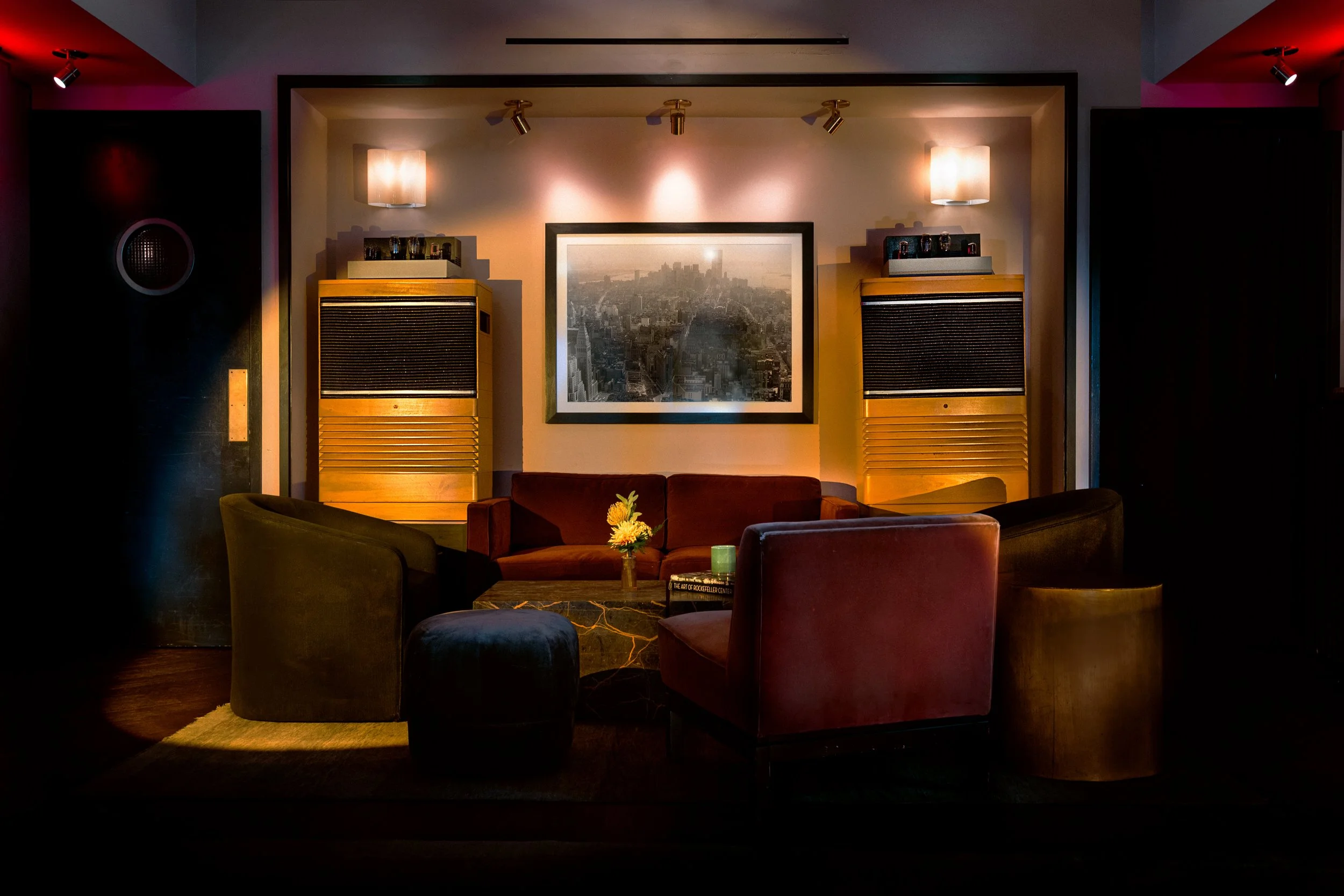 A cozy living room with a red sofa, green armchair, and round ottoman, centered around a black marble coffee table with a flower arrangement. Two large wooden speakers are mounted on the wall behind the sofa, with a framed black and white cityscape p