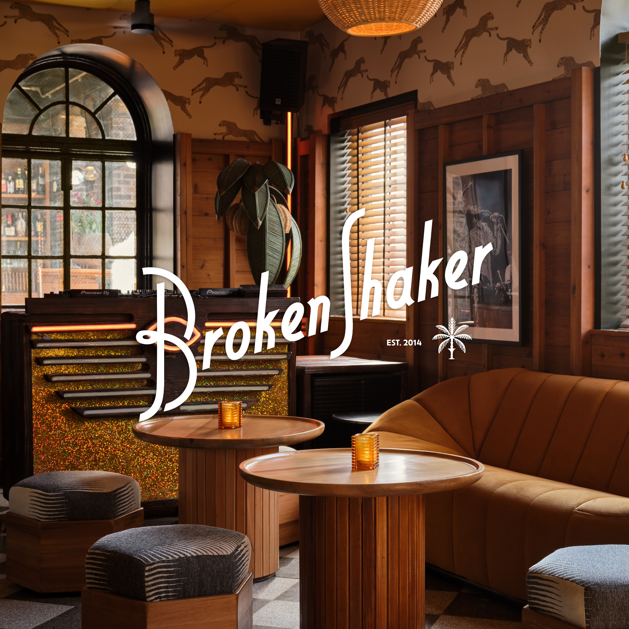 Interior of a cozy, wood-paneled bar or lounge featuring a large window, wooden tables with candles, a golden velvet sofa, framed pictures, and a DJ setup with a Pioneer mixer. The room has a warm, tropical vibe with leaf-patterned wallpaper and a hanging rattan light fixture.