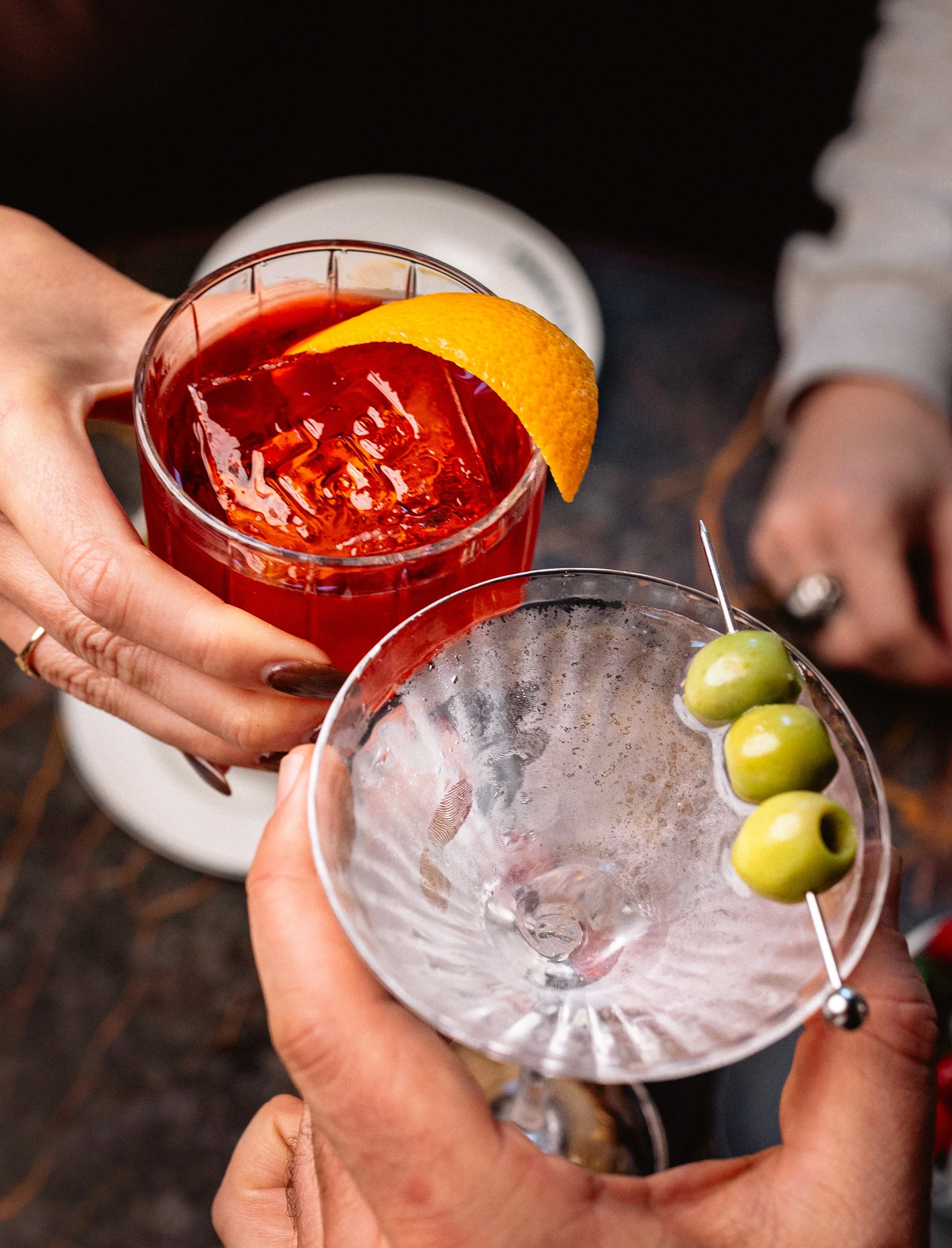 Two people clinking glasses of drinks, one with a red cocktail with an embossed "PB" ice cube and garnished with an orange peel and the other with a clear martini garnished with three green olives on a skewer.