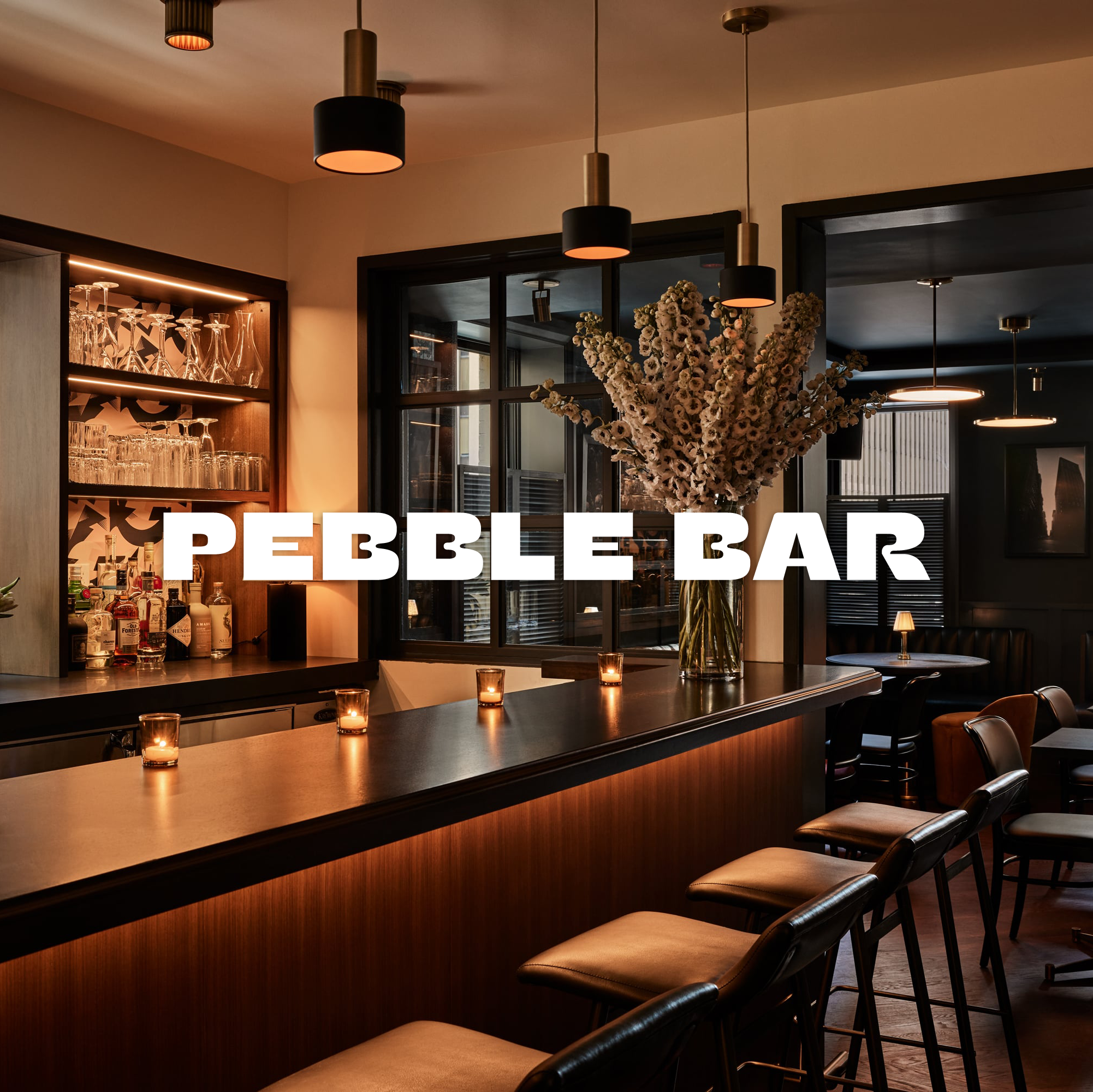Interior of Pebble Bar with bar counter, candles, large floral arrangement, shelves with glassware, and seating area with tables and chairs.