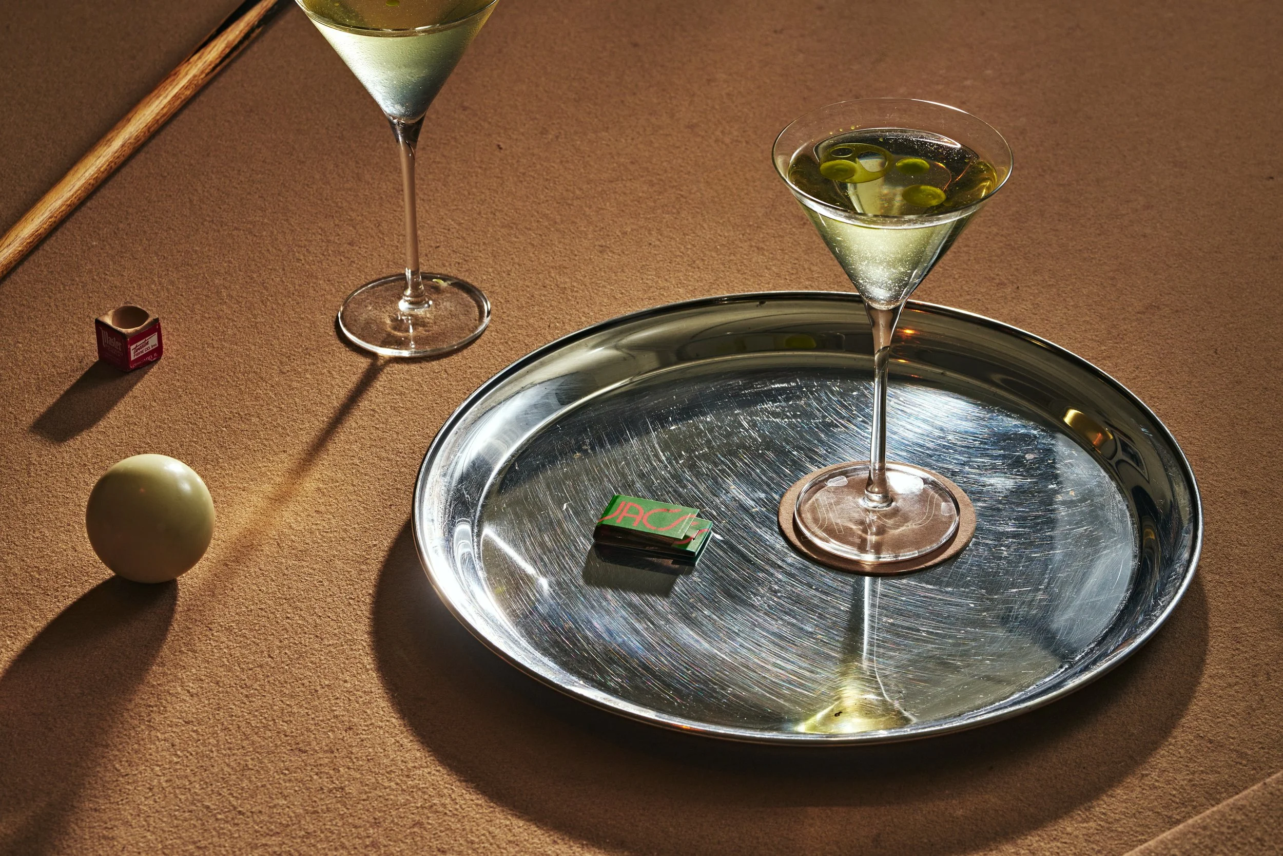 Two martini glasses filled with a clear cocktail and green olives, a silver tray, a pack of matches, a billiard ball, and a cue stick on a tan pool table.