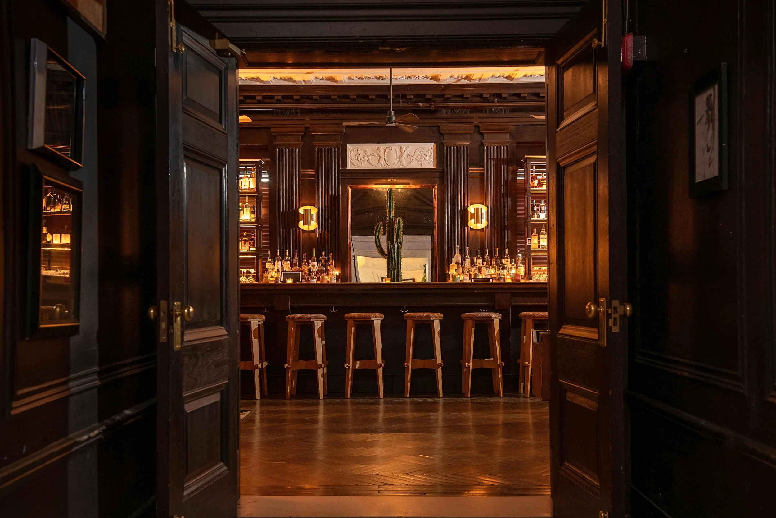 View of a dimly lit bar through dark wooden doors. The bar features a cactus in a black pot, a large mirror, and shelves with liquor bottles. The bar counter has five wooden stools.