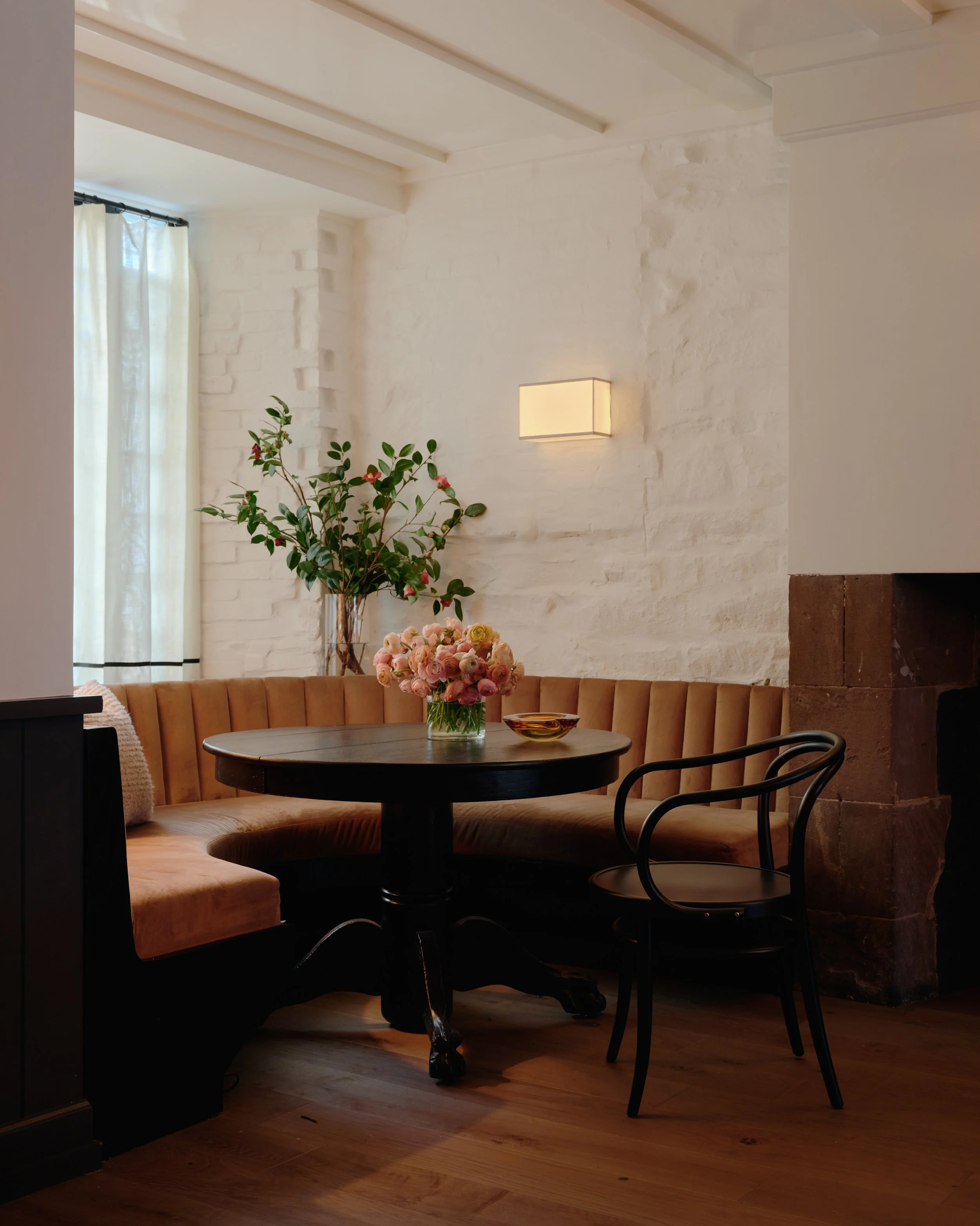 A cozy dining nook at Jac's on Bond with a round wooden table, pink flower bouquet, peach upholstered bench seat, black chair, green leafy plant, white brick wall, and a window with sheer curtains.