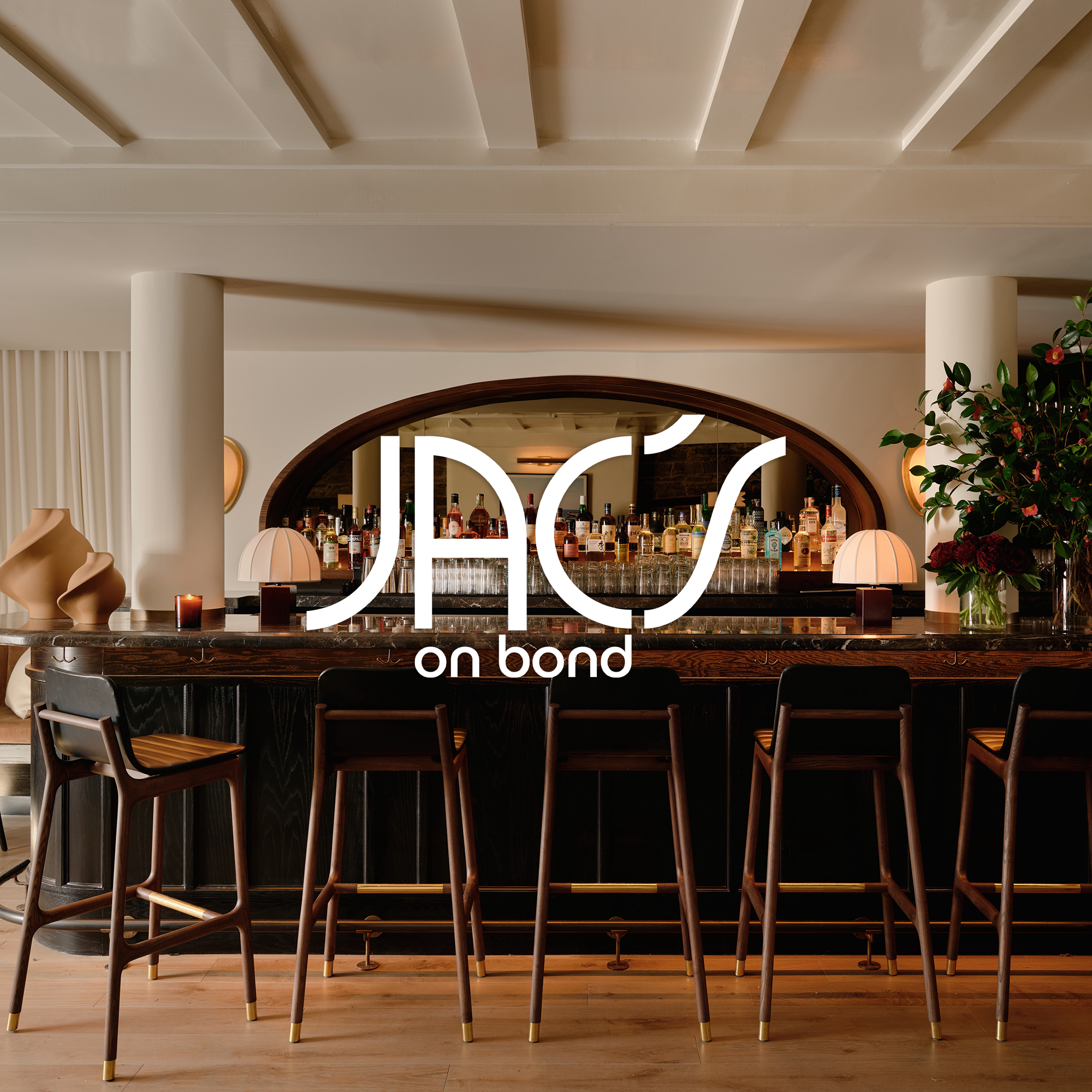 Modern bar with a dark wood counter, bar stools, bottles of alcohol on shelves behind, and decorative vases and lamps on the counter. Overlay text reads 'JAC on bond'.