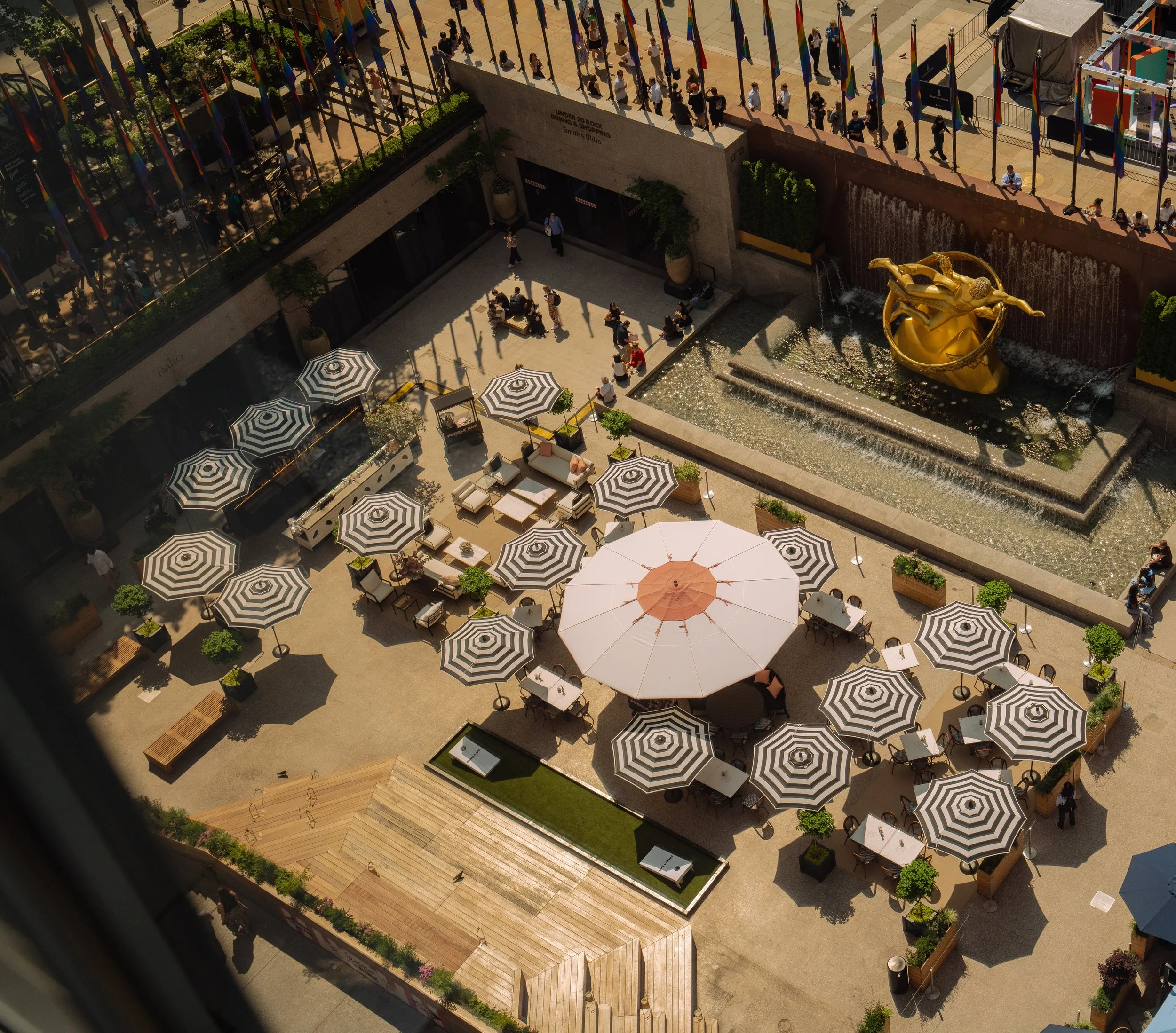 Aerial view of the outdoor seating area of Pebble Bar at the Rink at Rockefeller Center, with black and white striped umbrellas, a large pink and white umbrella, potted plants, and the historic fountain with golden Prometheus sculpture.