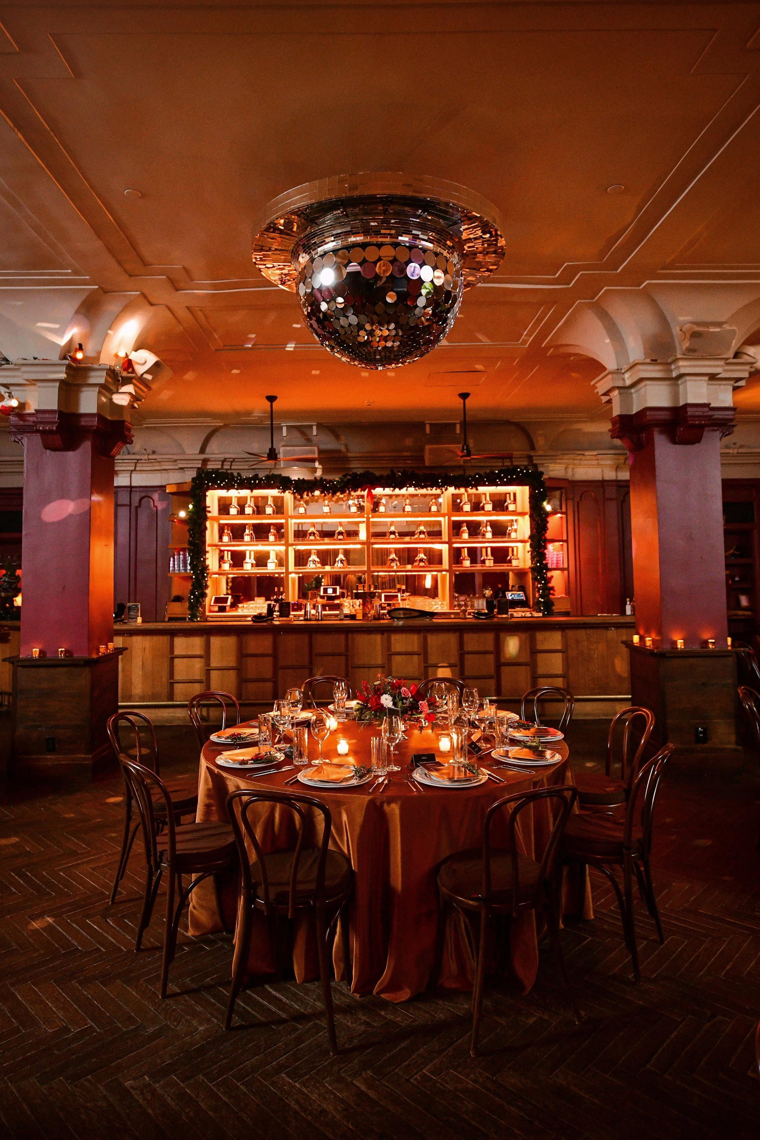Elegant banquet dinner table set with a floral centerpiece, candles, and place settings in the warmly lit Georgia Room event space with a bar in the background and a large mirrorball on the ceiling.