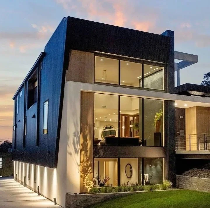 Modern multi-story house with large glass windows, illuminated interior, and a landscaped front yard at sunset.