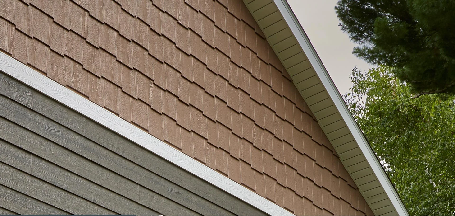 Close-up of the exterior of a house showing brown shingle siding and gray horizontal siding, with a white trim separating them, and part of a roof with trees in the background.