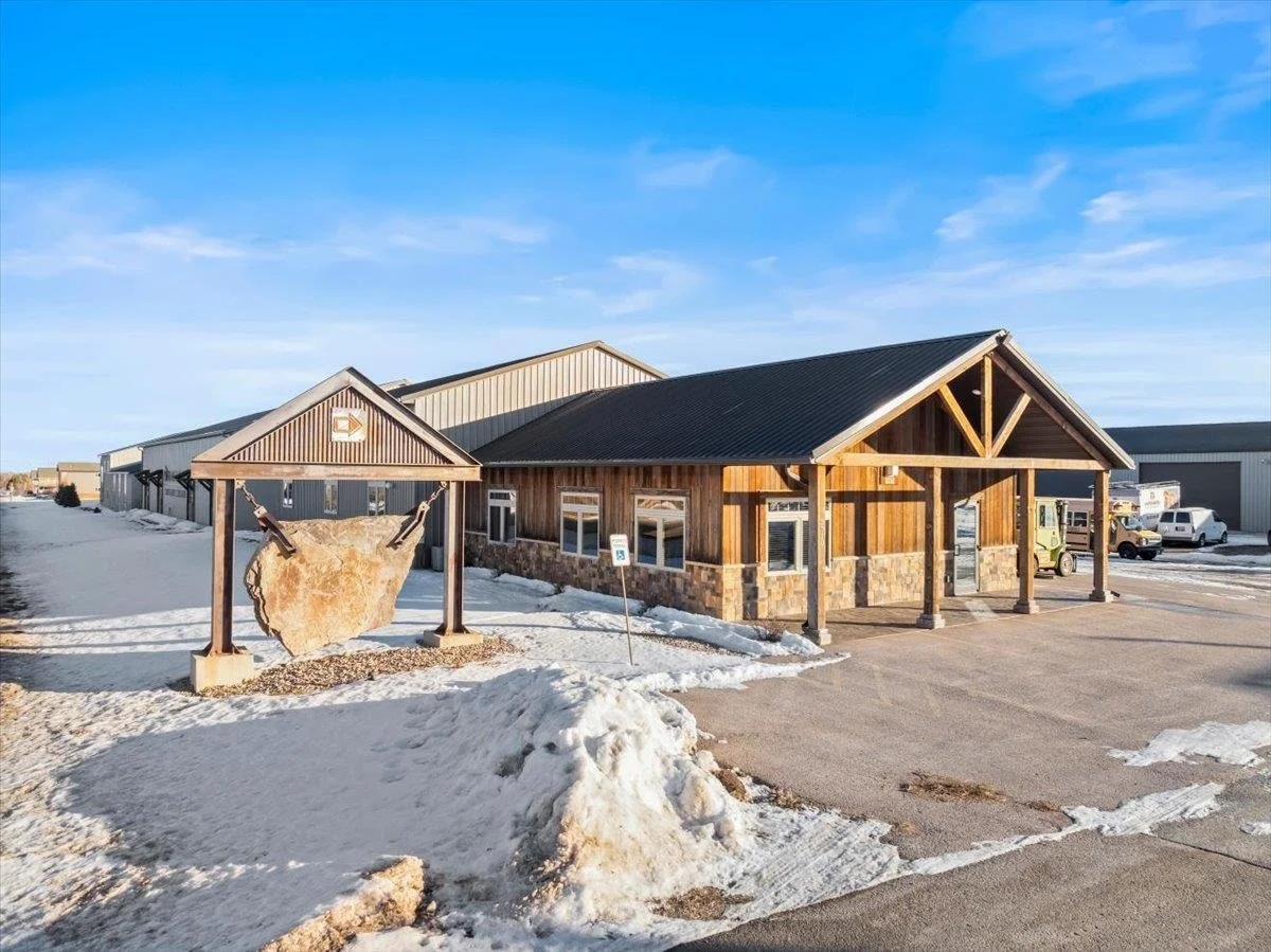 A building with wooden siding and stone accents, situated in a snowy landscape under a blue sky, with a large stone suspended by chains outside.