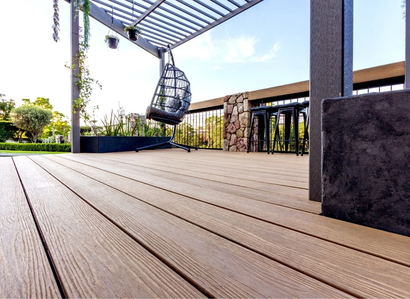 A wooden porch with a black hanging chair, potted plants, and a bar stool set under a pergola against a blue sky with trees in the background.