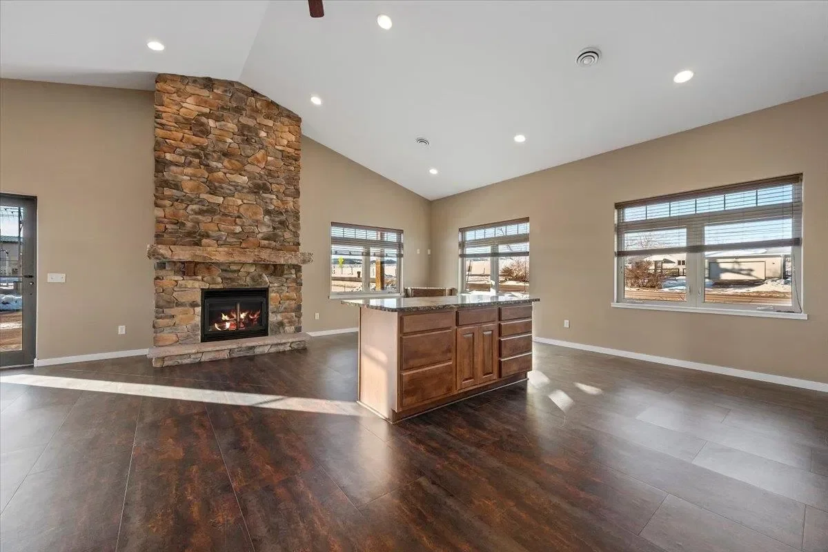 Spacious living room with a stone fireplace, hardwood floors, three windows, and a kitchen island with wooden cabinets.