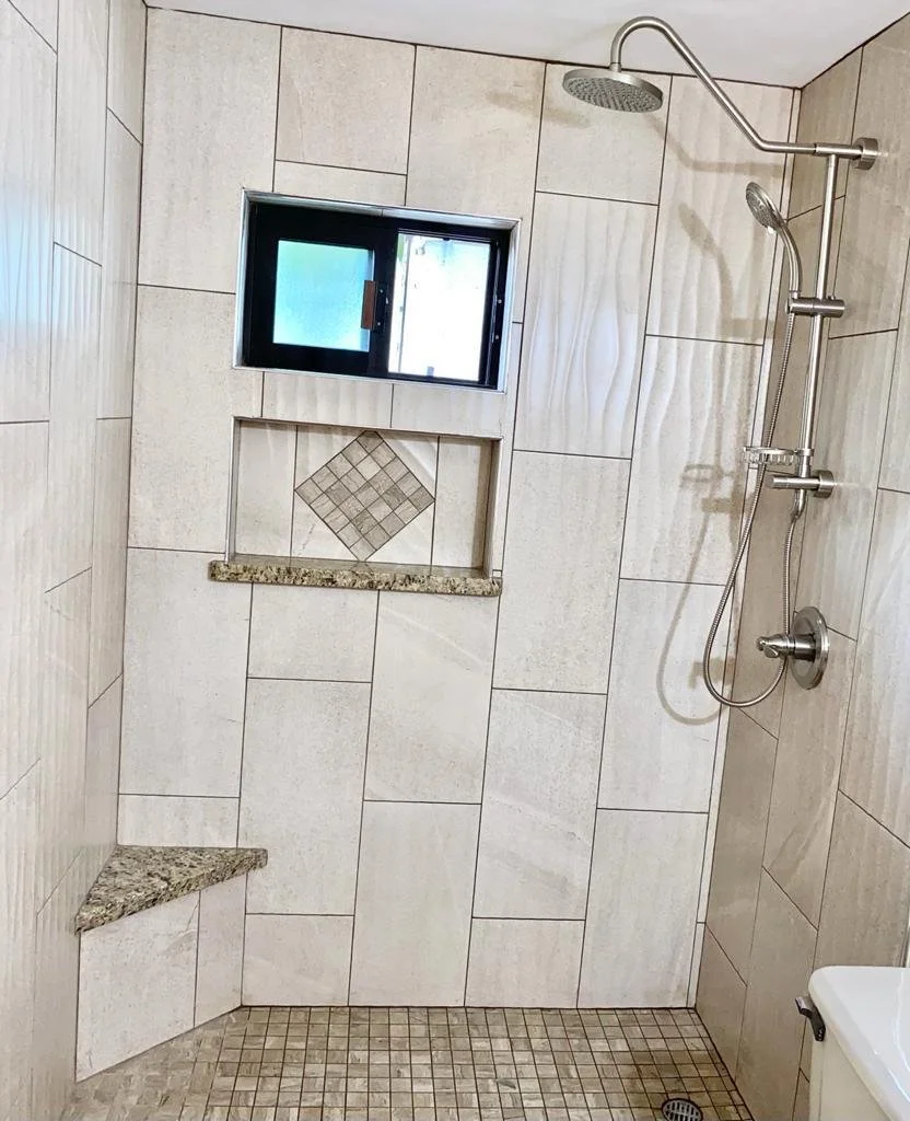 A shower area with beige tile walls, a small window, built-in granite niche, and bench with granite top. Stainless steel shower fixtures and a small square drain on the tiled floor.