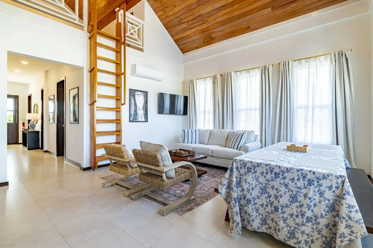 Bright living room with high wooden ceiling, white walls, and large windows with curtains. Contains a white sofa with striped pillows, a wooden coffee table, two woven chairs, and a flat-screen TV on the wall.