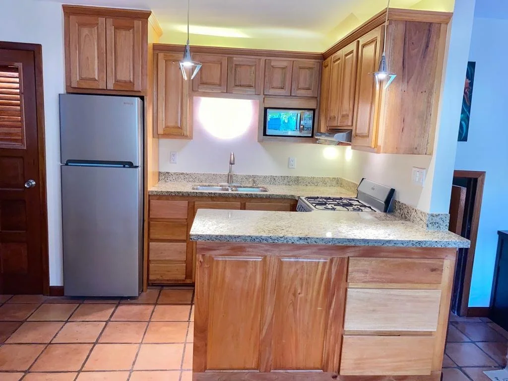 Kitchen with wooden cabinets, a granite countertop, stainless steel refrigerator, microwave, gas stove, tile flooring, and pendant lighting.
