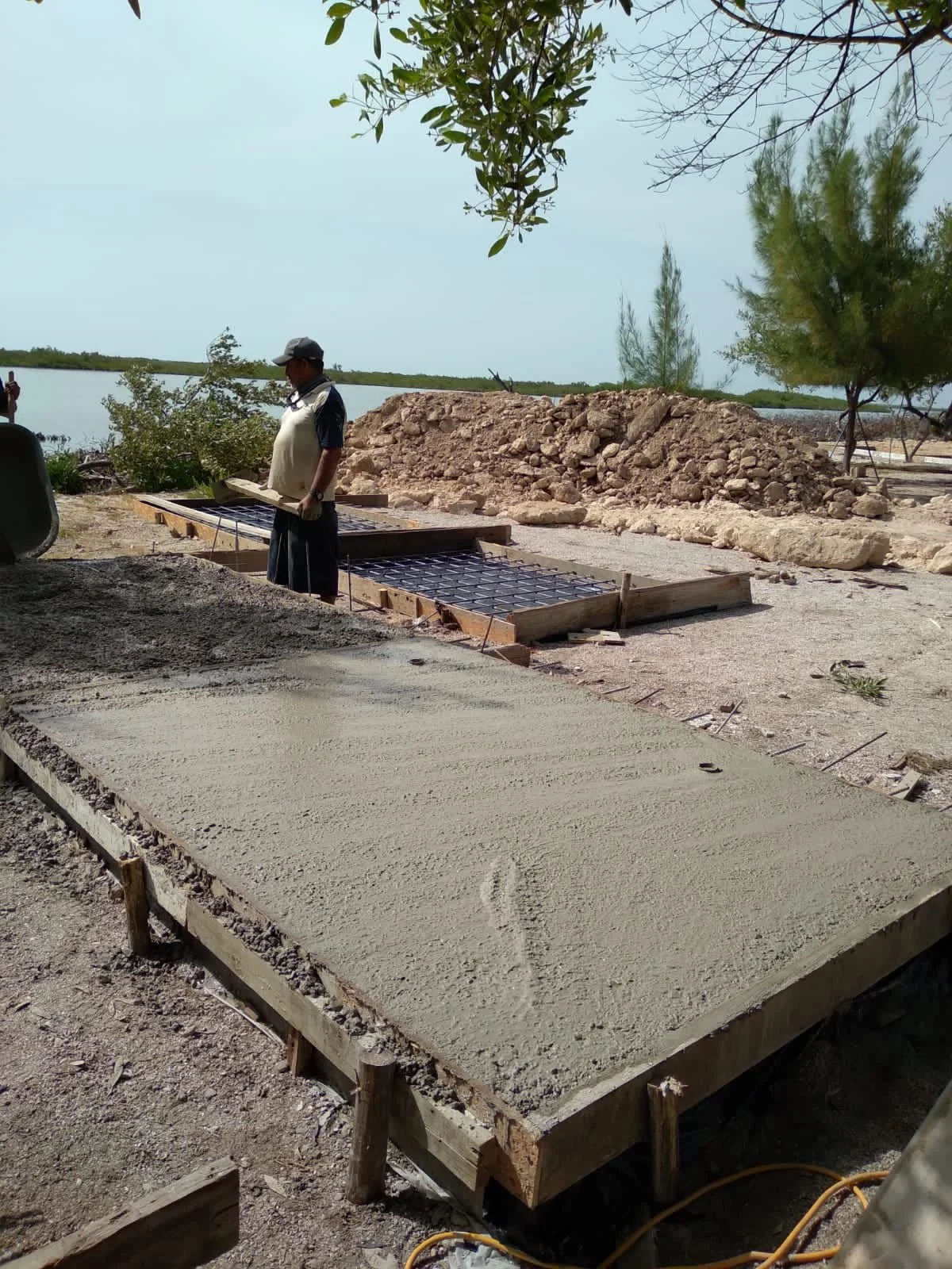 Construction site near water with a worker pouring and smoothing concrete in molds, surrounded by dirt, trees, and rocks.
