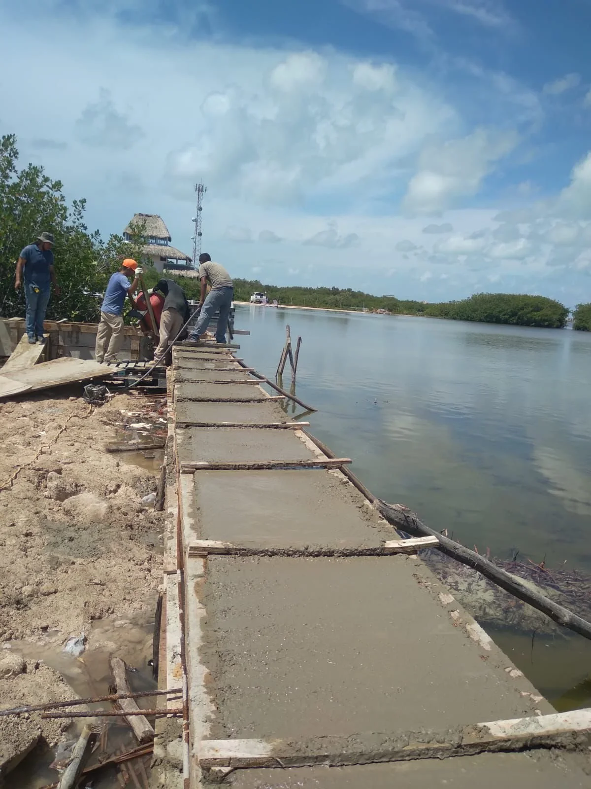 Construction workers building a concrete walkway along a waterway on a partly cloudy day.