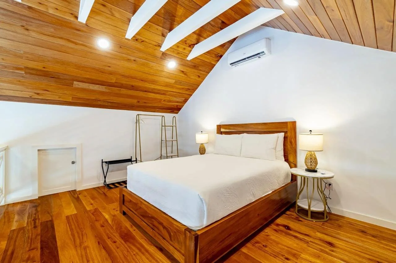 A bedroom with a wooden floor and ceiling, white walls, a large bed with white bedding, two matching lamps on side tables, and decorative ladders in the corner.