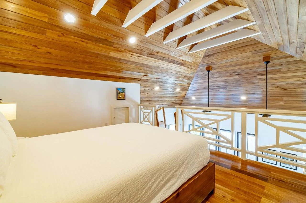 A bedroom with a white bed and wooden flooring, featuring a loft style with wooden paneling on the ceiling and white railings along the edge of the loft.