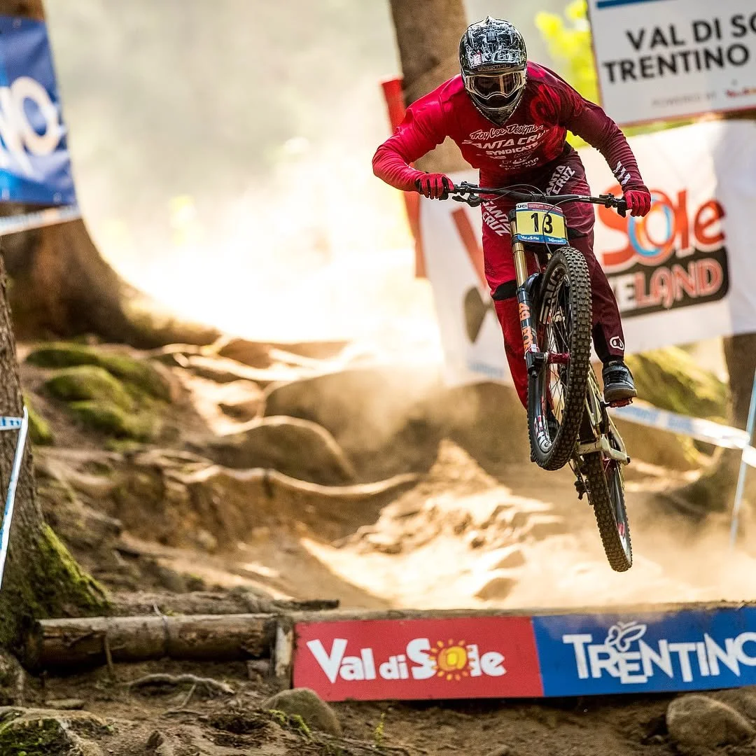 Mountain biker wearing red gear and helmet jumps off a dirt trail on a forested downhill course, with banners and signs in the background.