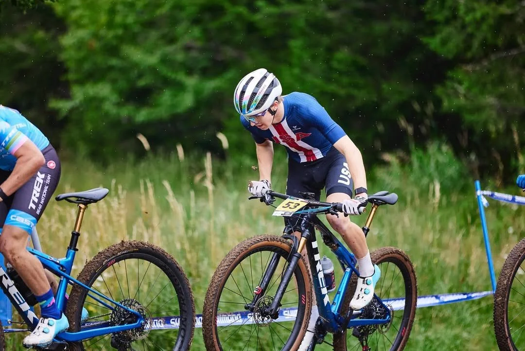 A young male cyclist in a blue USA uniform and helmet, riding a mountain bike with muddy tires, during a race on a grassy trail with trees in the background.