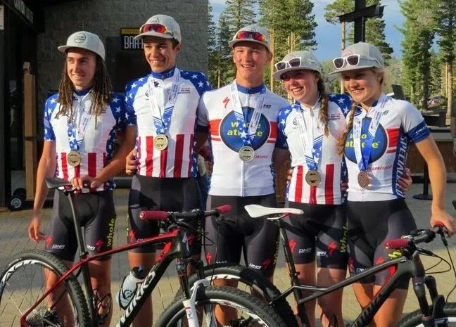 Five young cyclists wearing American flag-themed jerseys and medals around their necks, standing outdoors with bicycles, in front of a wooded background.