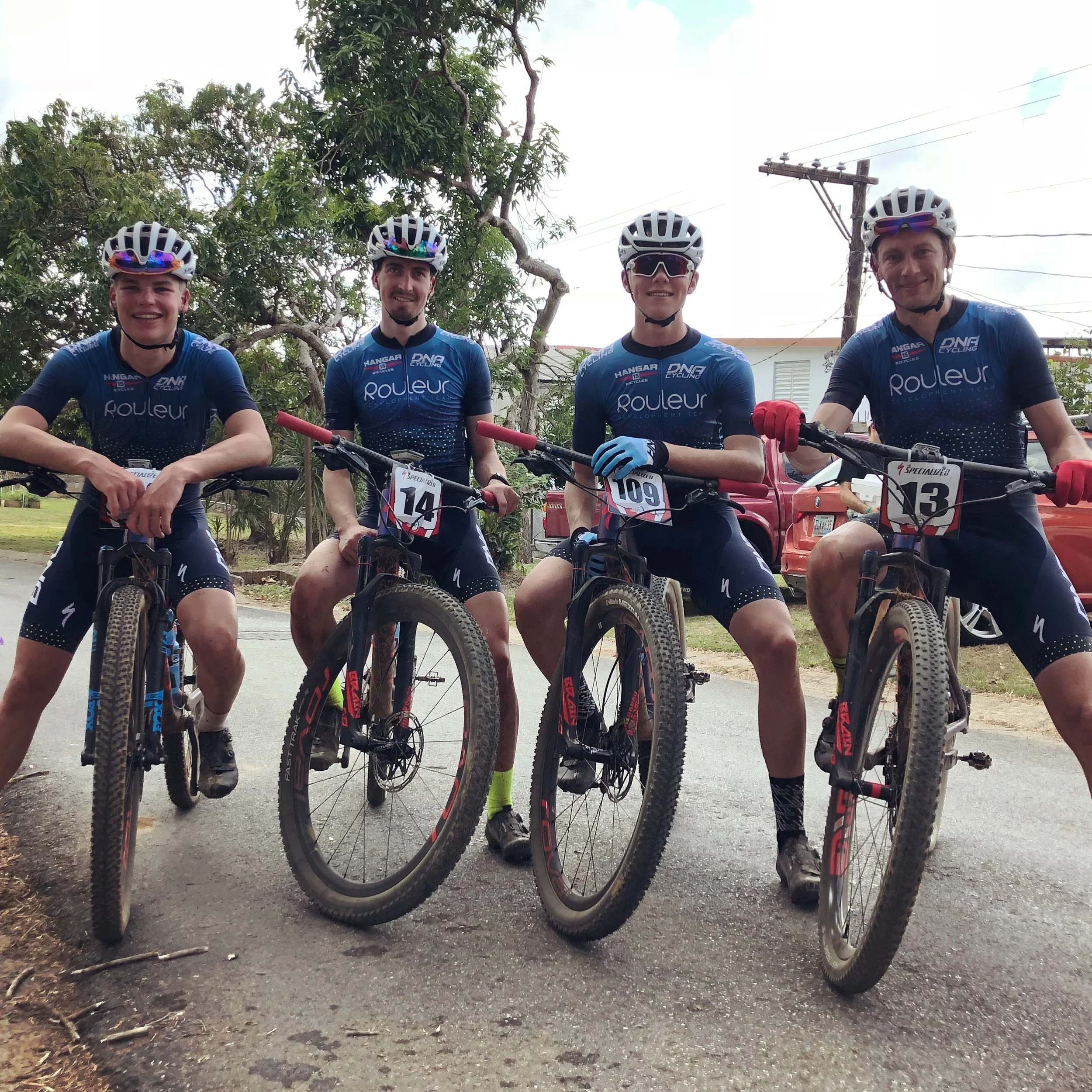 Four male cyclists in matching blue jerseys and helmets, sitting on mountain bikes, posing outdoors with trees and utility poles in the background.