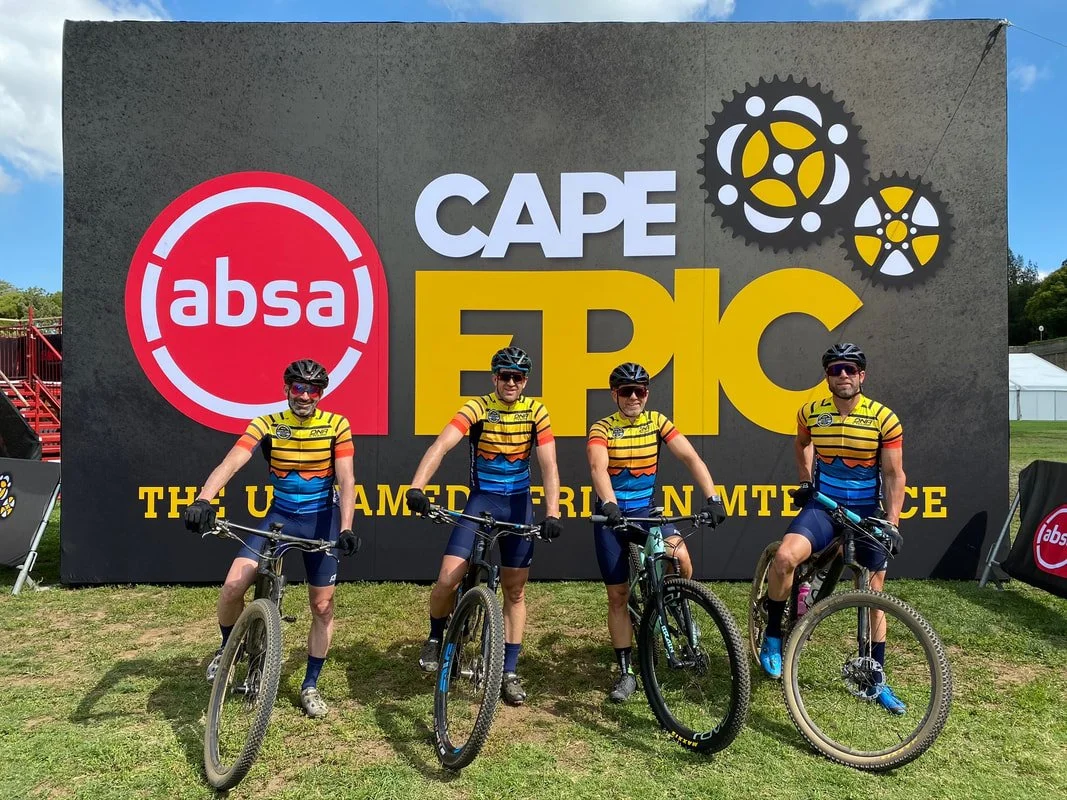 Four cyclists in matching yellow, blue, and orange jerseys pose with their bikes in front of a large sign that reads "Cape Epic," with a red "absa" logo on the left and gear imagery on the top right. The event is labeled as "The Ultimate Mountain Race."
