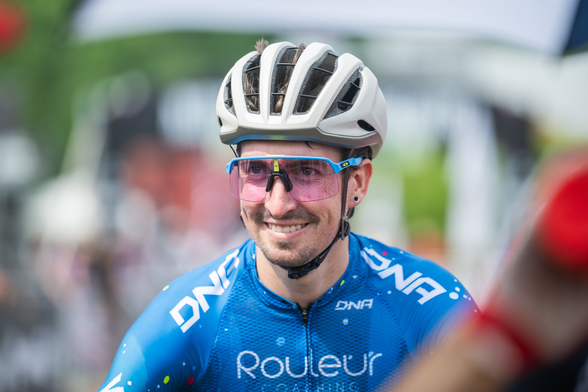 A male cyclist wearing a white helmet, blue sunglasses, and a blue cycling jersey smiling at an outdoor event.
