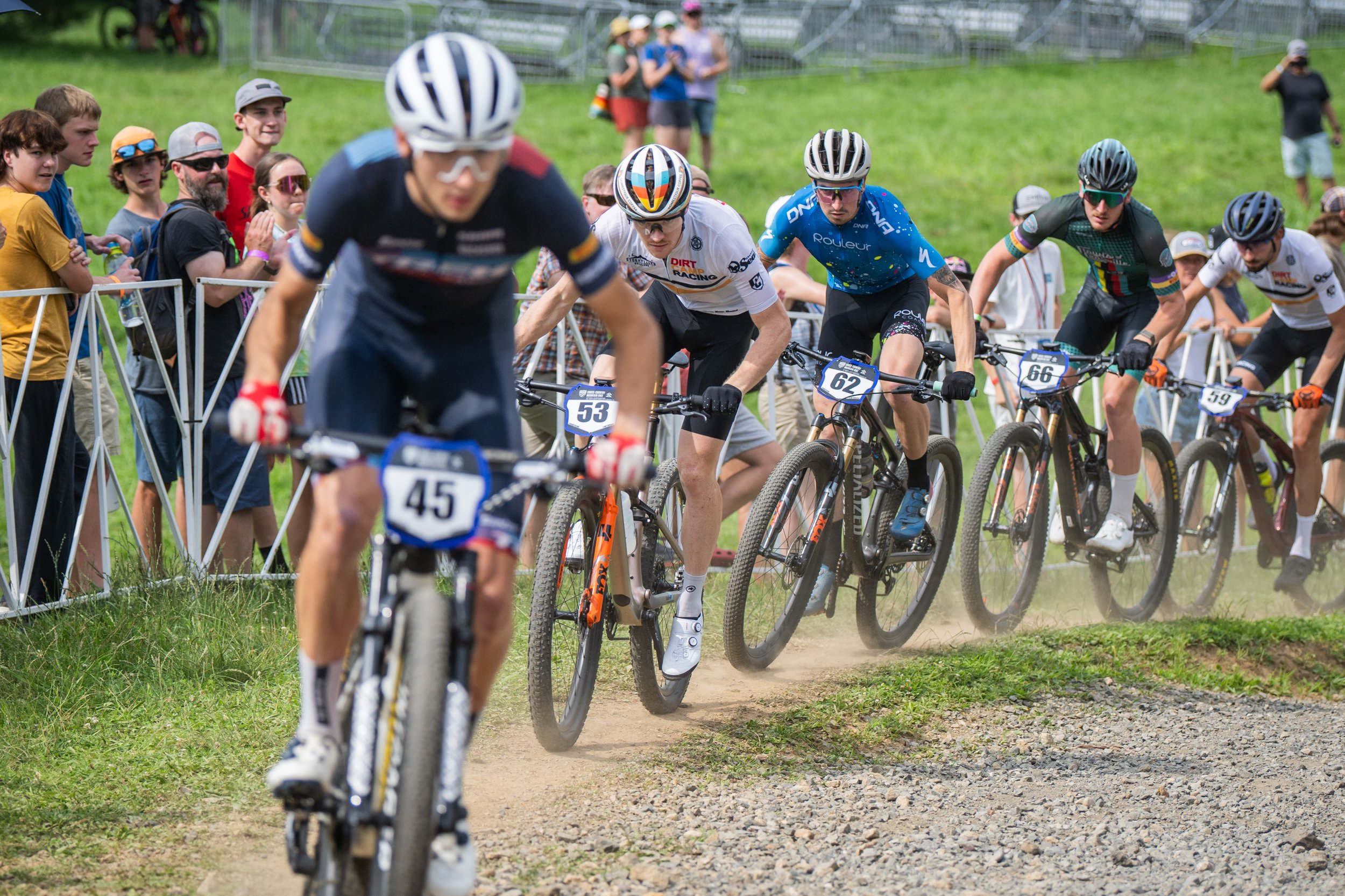 Group of mountain bikers racing on a dirt trail, with spectators watching behind a barrier.