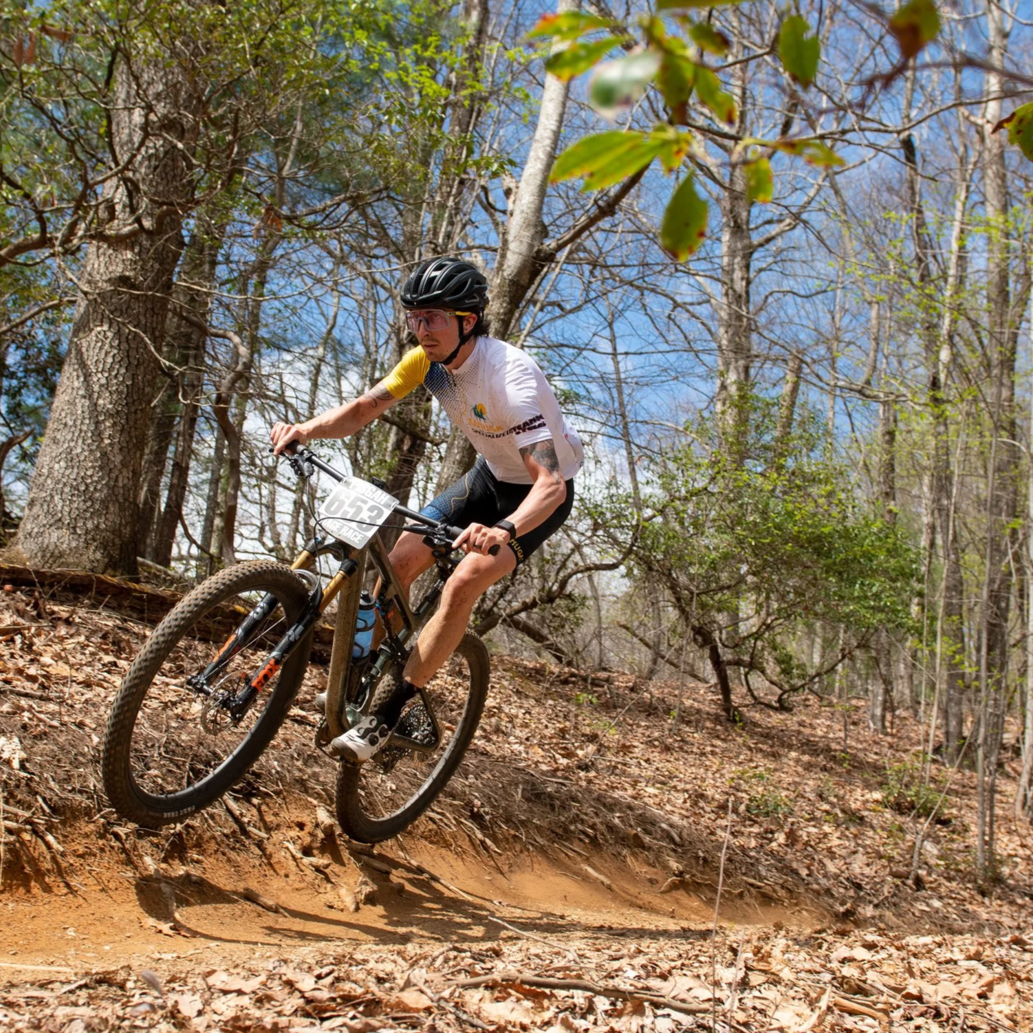 A mountain biker wearing a black helmet, sunglasses, and a white jersey with yellow sleeves riding on a dirt trail through a wooded area with trees and leaf litter on the ground.