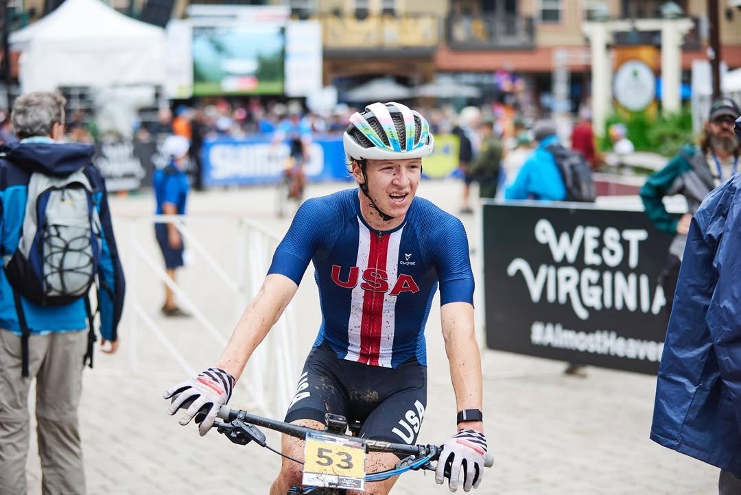 Female cyclist with USA jersey and helmet, smiling, holding bike handlebar, at an outdoor race with people and signs in West Virginia background.