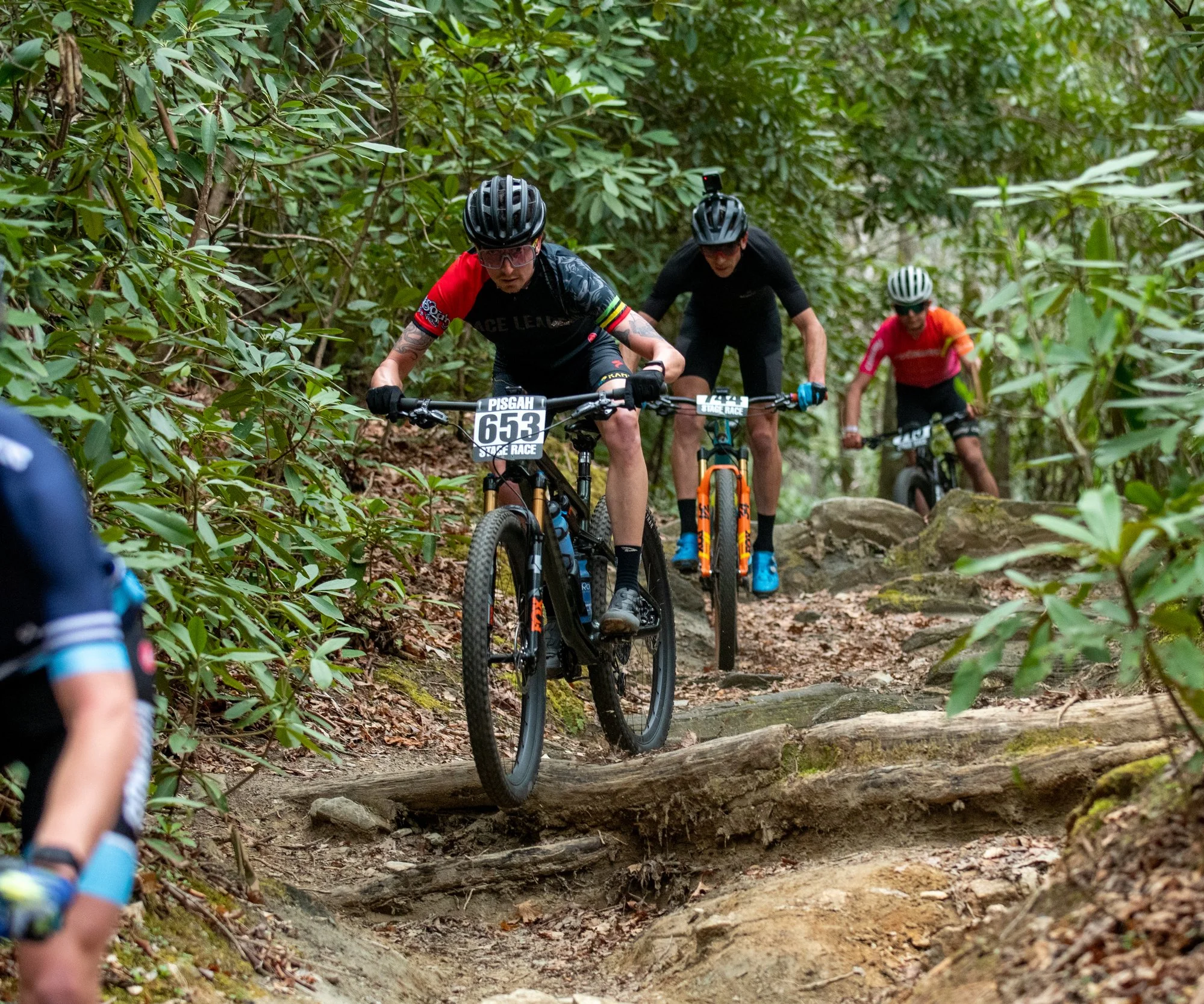 Four mountain bikers riding on a dirt trail through a forest, with one rider in the front jumping over a log and others following behind.