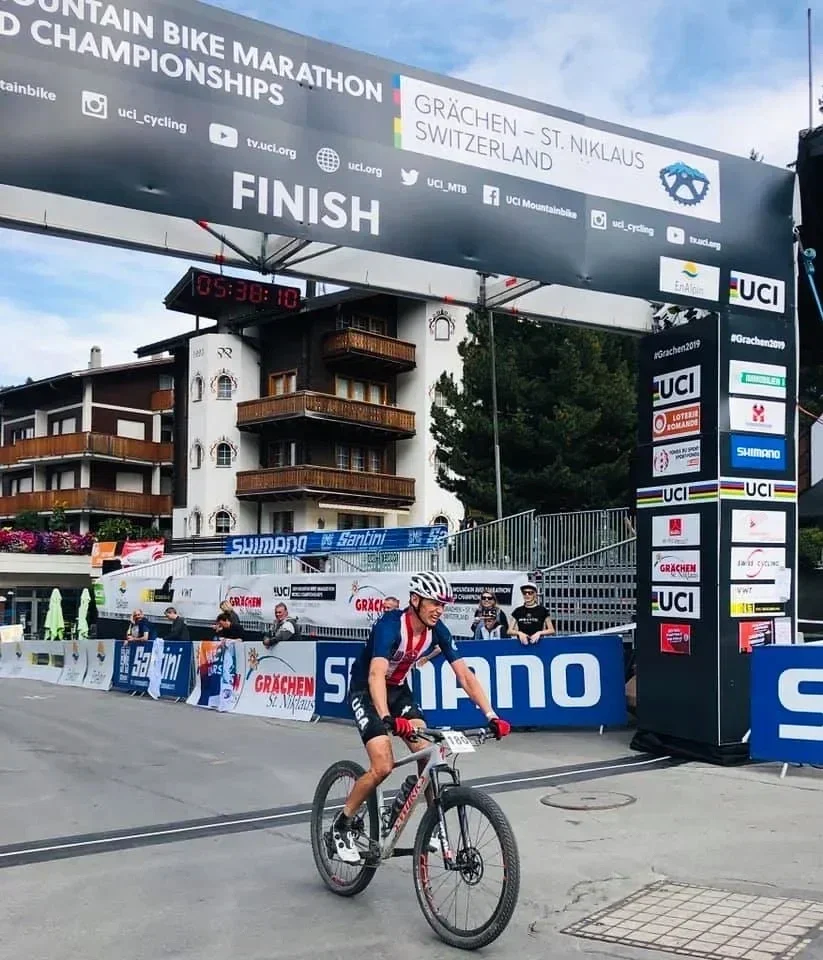 Cyclist crossing the finish line at a mountain bike marathon race in Grachen, Switzerland, with a large banner overhead reading 'UCI Mountain Bike Marathon World Championships' and a digital timer showing 5 hours, 38 minutes, and 10 seconds.