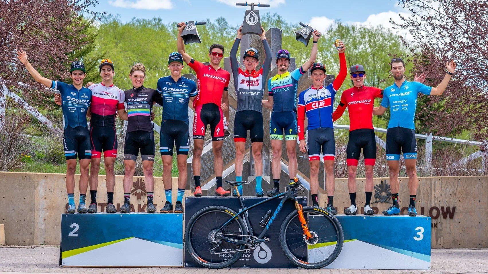 Cyclists on a podium celebrating victory, with some holding trophies and a mountain bike in front, set outdoors with greenery in the background.