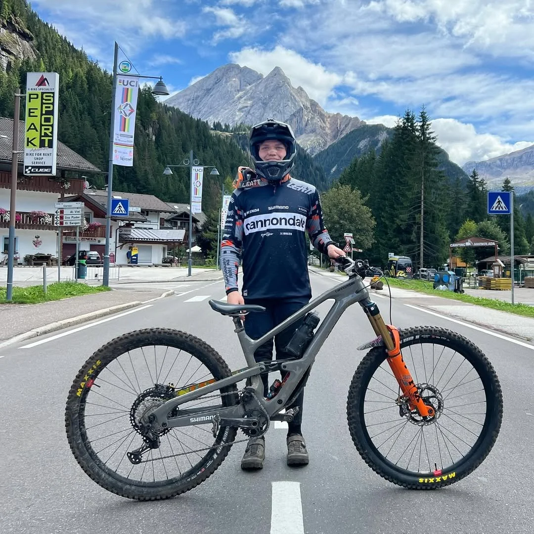 A person dressed in cycling gear standing with a mountain bike on a paved road in a mountain town, with tall mountains and a partly cloudy sky in the background.