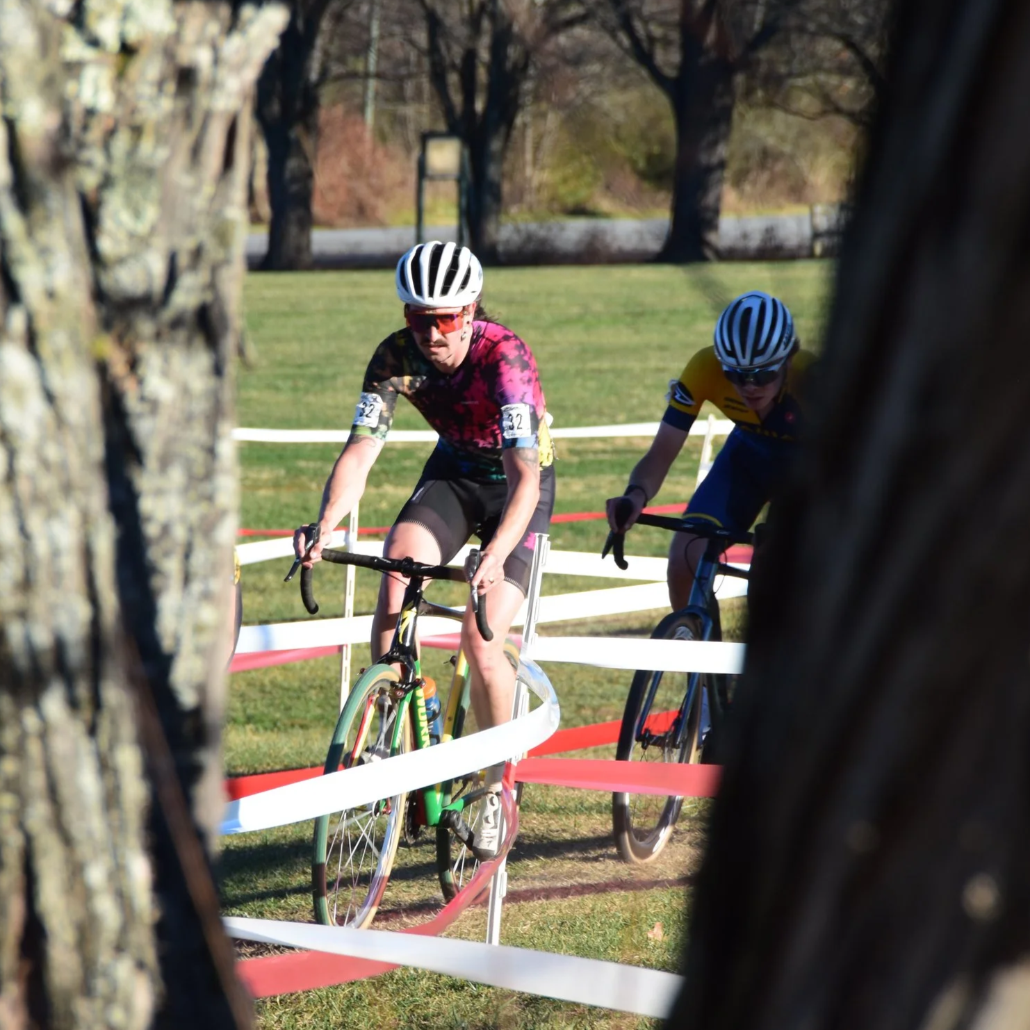 Two male cyclists wearing helmets and sports gear navigate a cyclocross racecourse lined with red and white barriers, seen through the gaps between trees in a park with green grass and trees in the background.