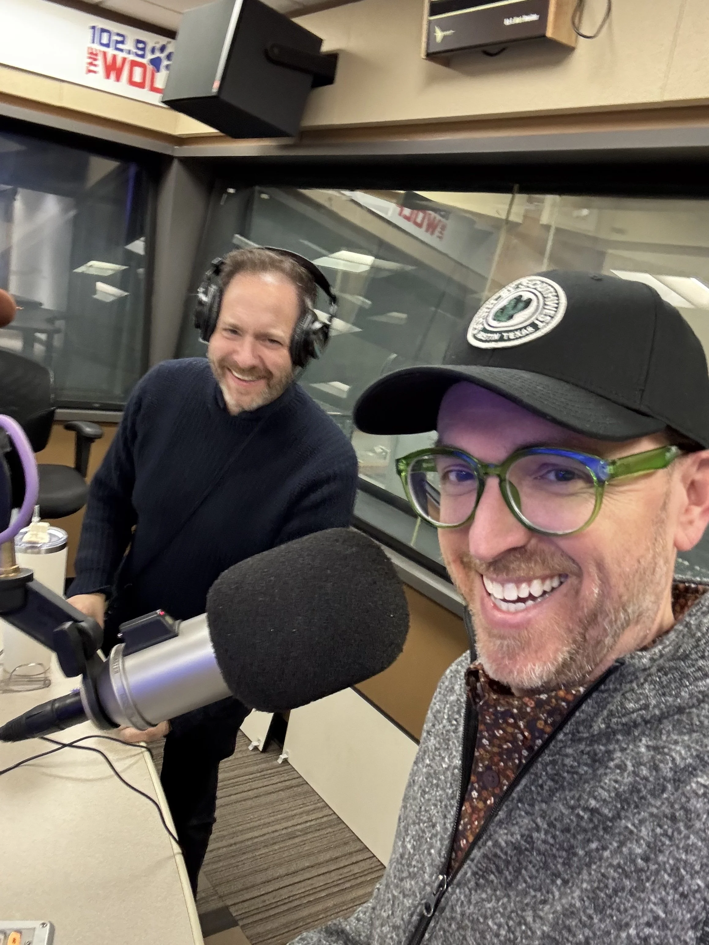 Two men smiling in a radio station studio, wearing headphones and speaking into a microphone, with radio station equipment and large windows in the background.