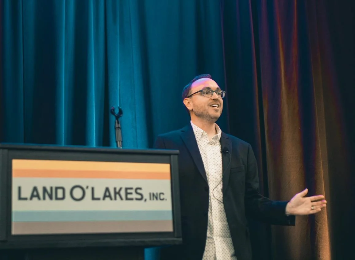 A man giving a presentation at a podium labeled 'Land O'Lakes, Inc.' with a dark blue curtain backdrop.